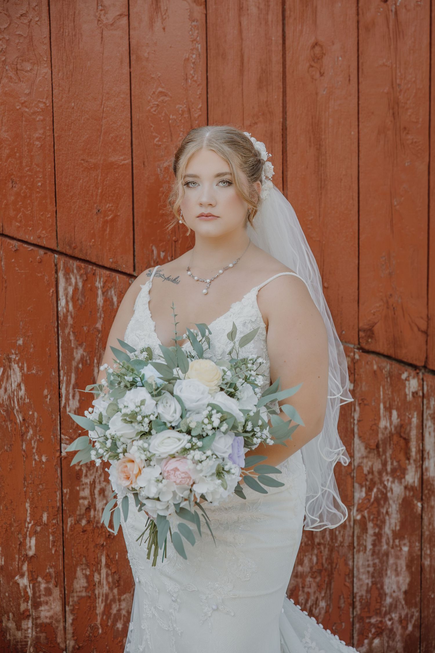 Bride in white lace mermaid gown holding floral bouquet, standing before rustic red barn wall.