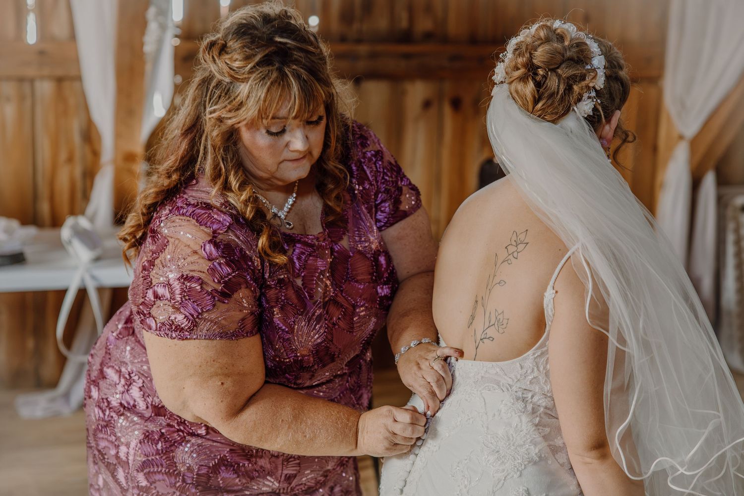 Mother helps bride with wedding dress buttons in rustic barn before ceremony.