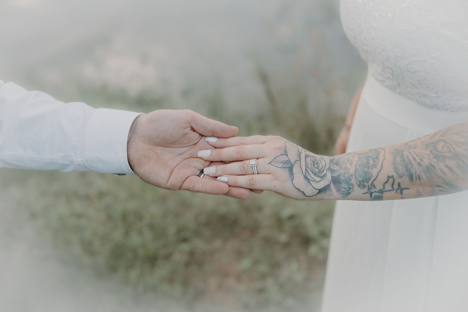Bride and groom holding hands showing wedding rings, with rose tattoo on brides arm, soft bokeh background.