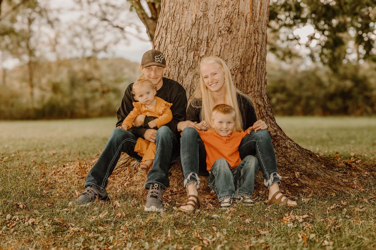 A group sits together on fallen autumn leaves under a large tree during a golden hour photo session.
