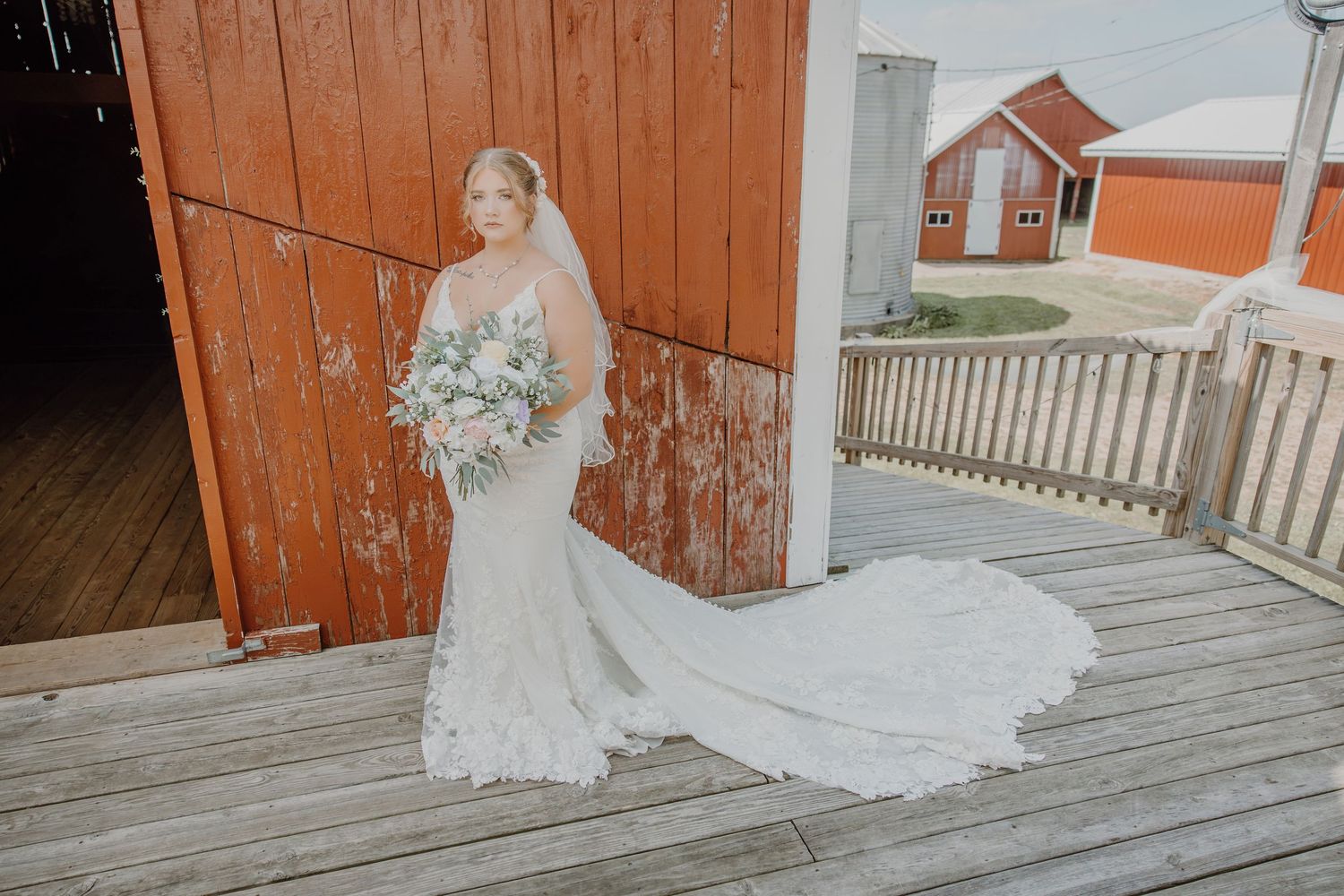 Bride in lace mermaid gown with veil and bouquet standing on wooden deck near rustic red barn.