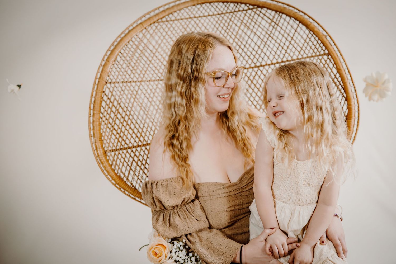 A woman with red curly hair and glasses sits with a young blonde girl in a wicker peacock chair, both smiling warmly.