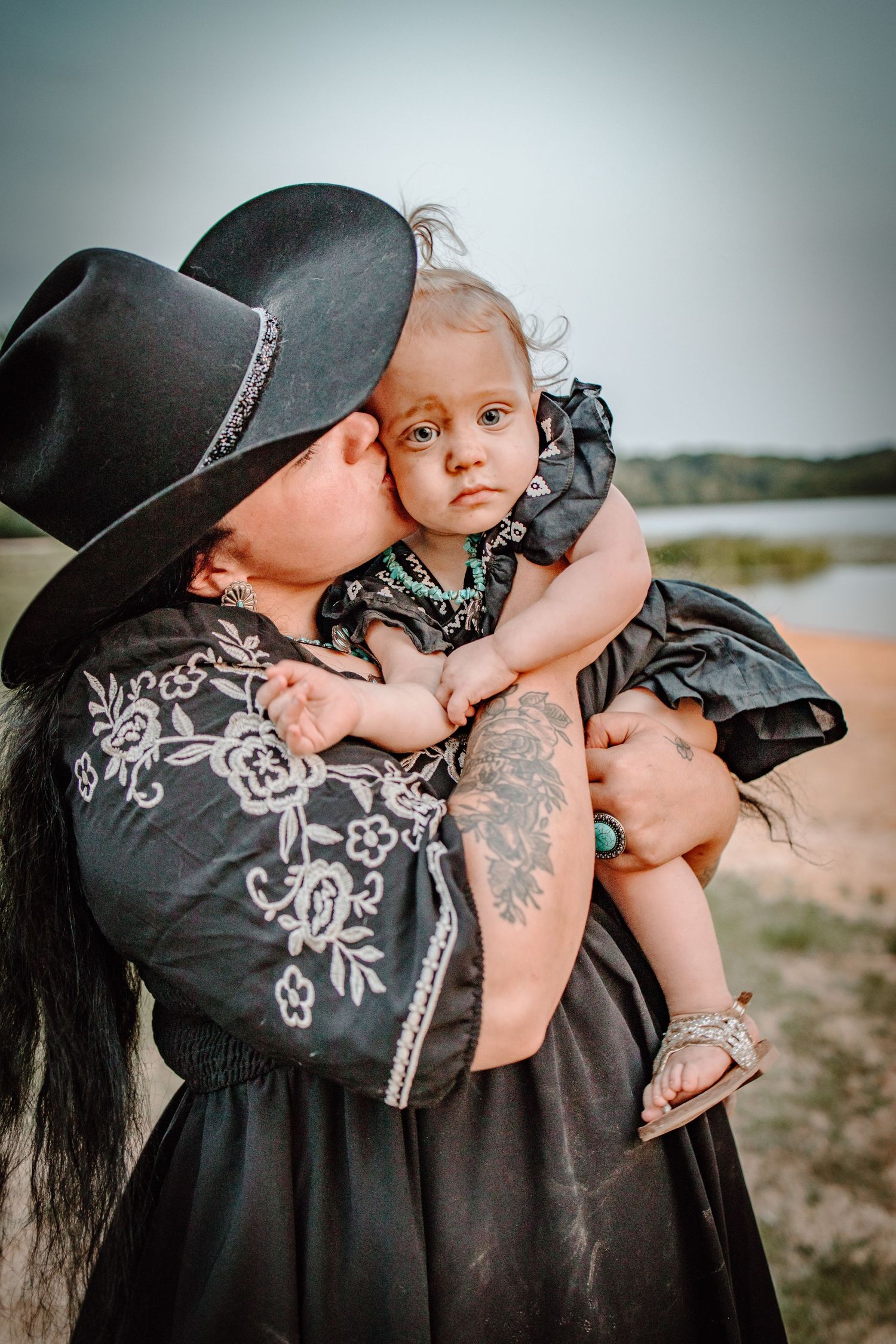 Parent in black embroidered dress holding and kissing small baby in outdoor setting at sunset.