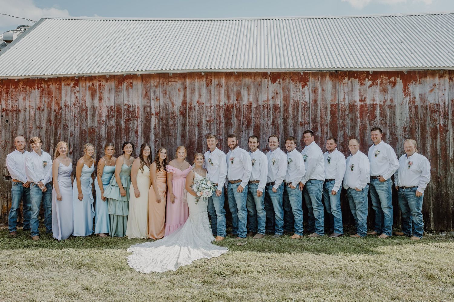 Wedding party poses before rustic barn, bridesmaids in pastel gowns and groomsmen in white shirts and jeans.