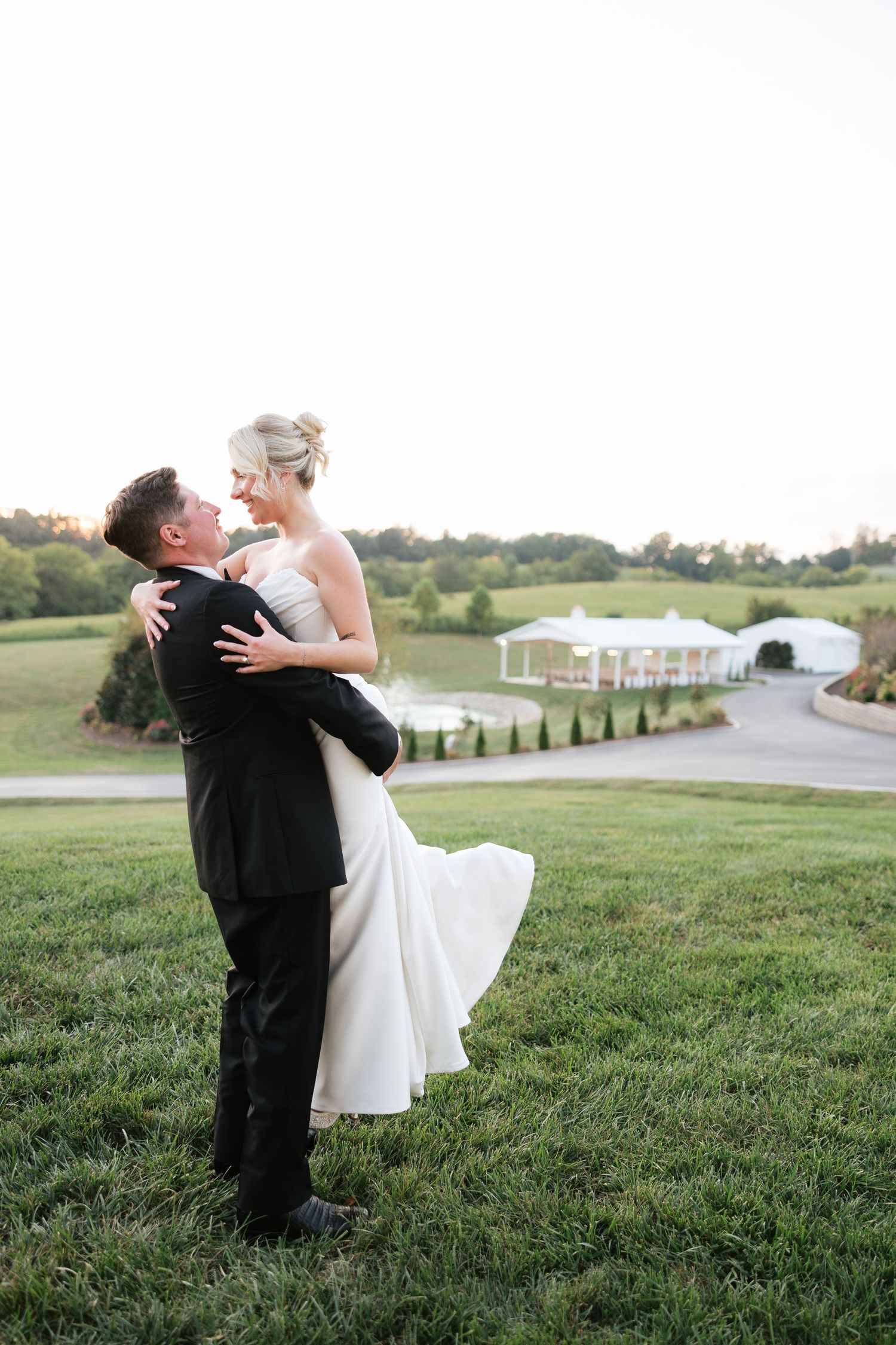 Groom lifts bride in white gown on green hillside with elegant wedding venue in background at sunset.