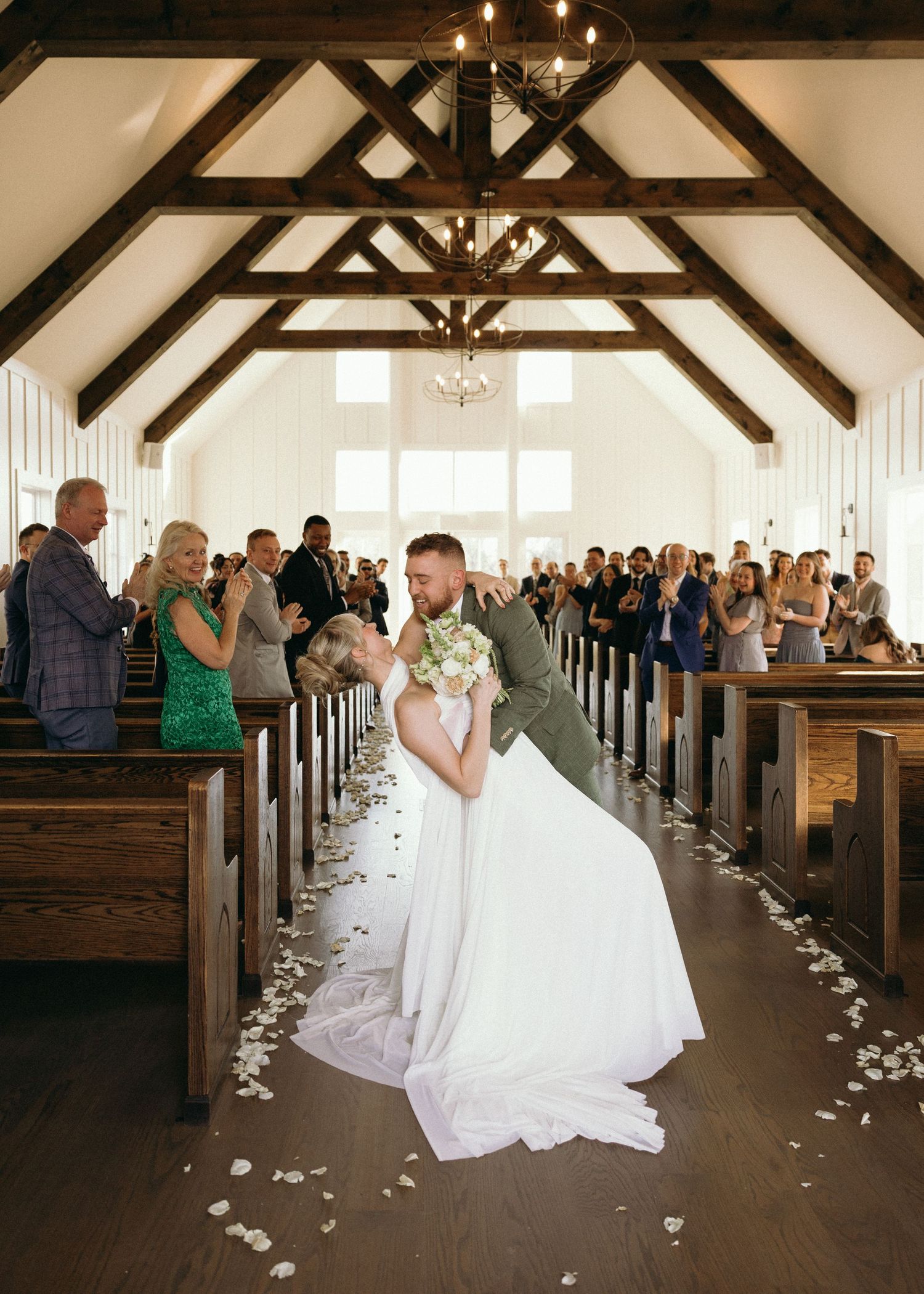 Bride and groom share a romantic dip kiss in a white chapel aisle surrounded by cheering wedding guests.