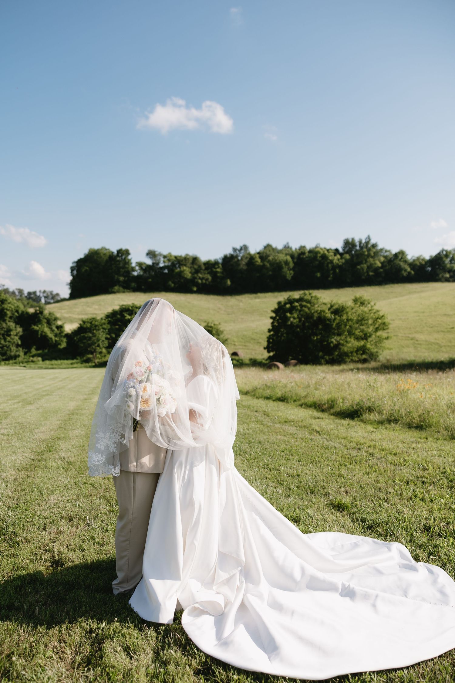 Bride and groom embracing under a flowing white veil in a lush green countryside field on a sunny wedding day.