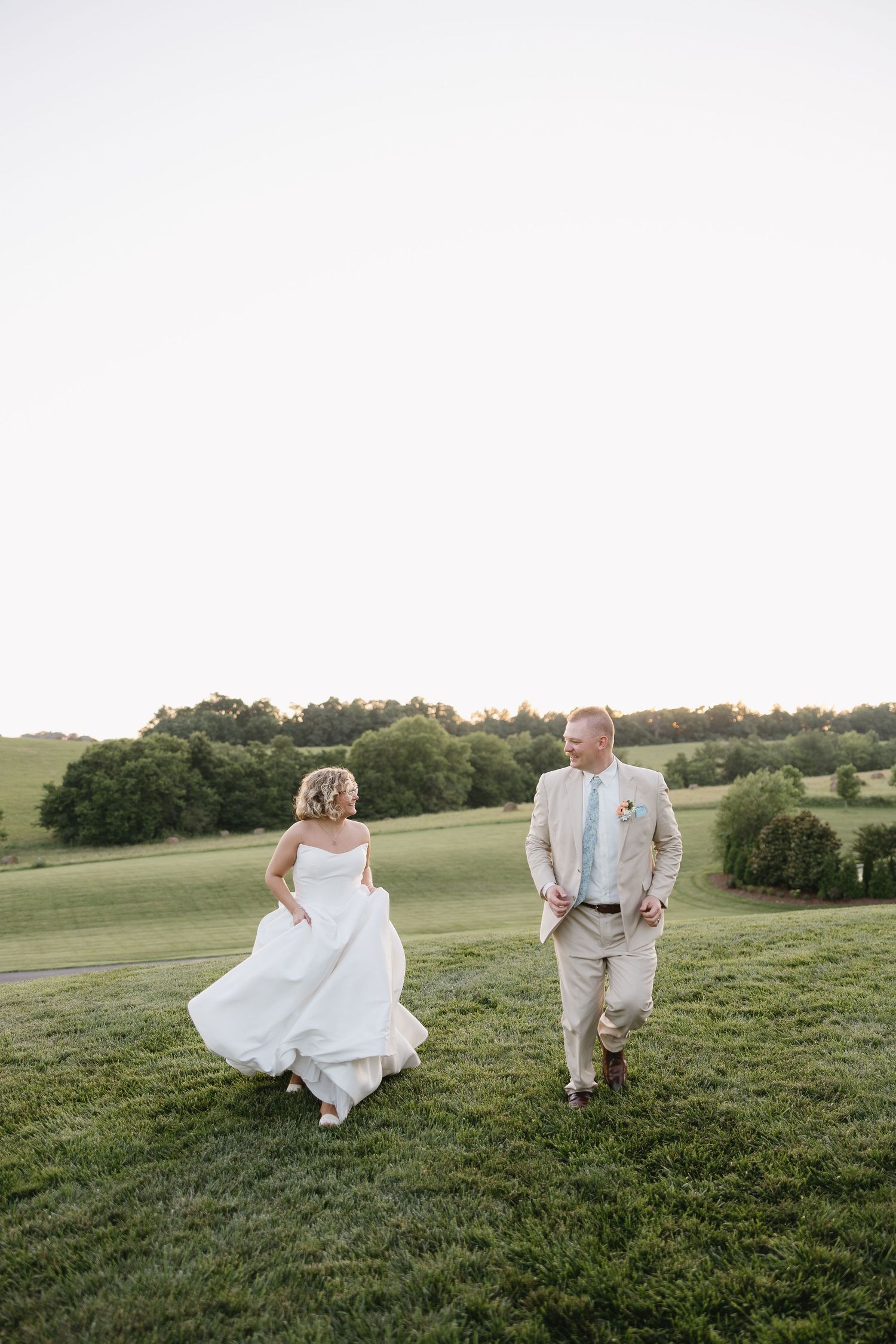 Bride in white gown and groom in tan suit walk hand in hand across lush green field at golden hour.