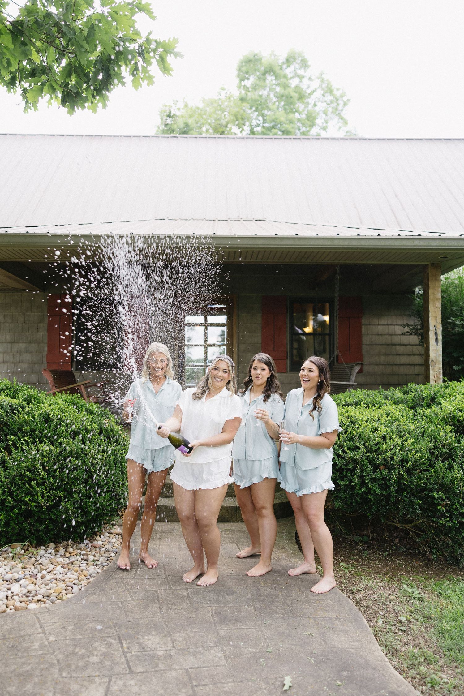 Four women in matching pajamas celebrate with champagne spray outside a rustic home.