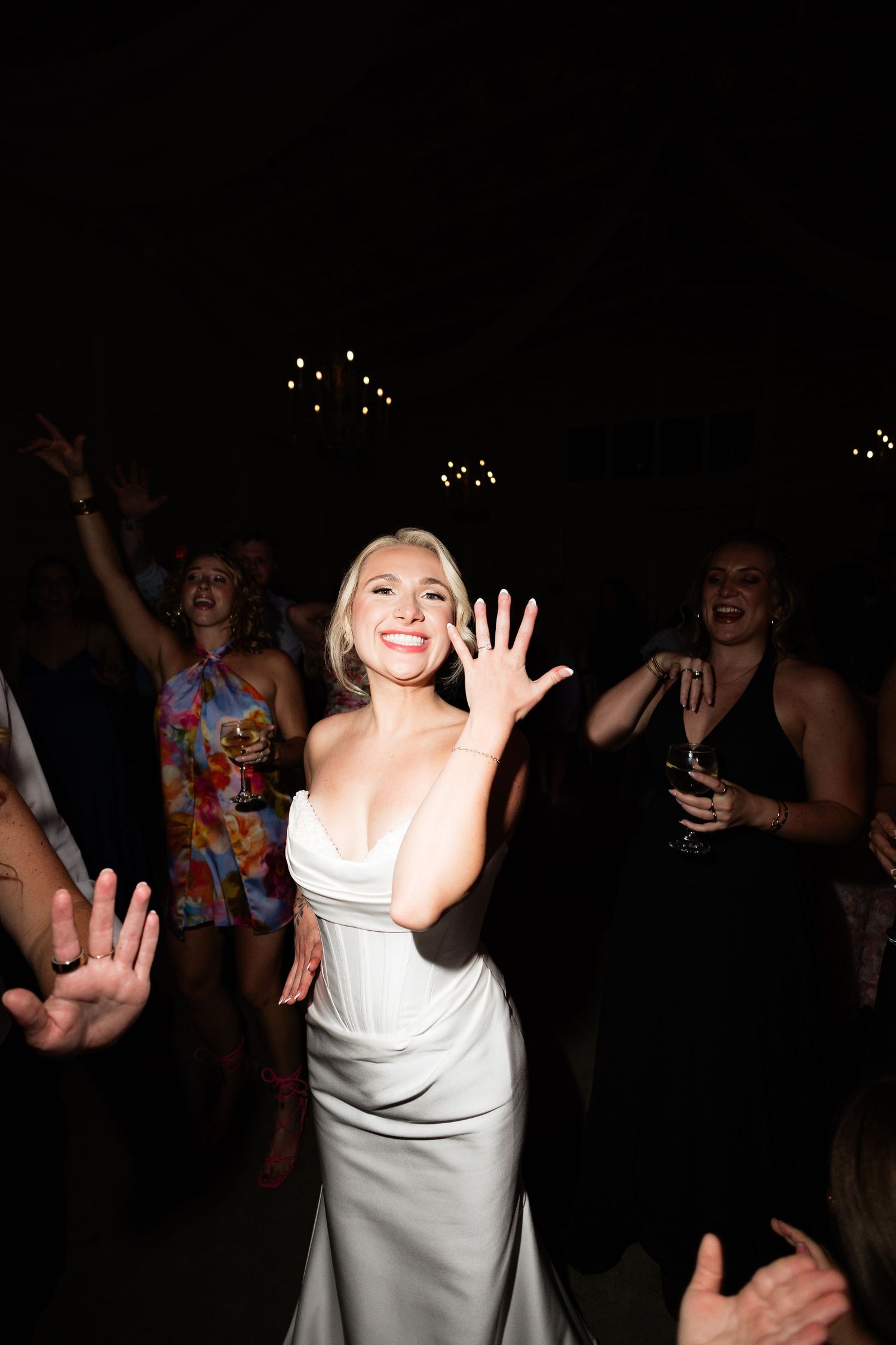 A radiant bride in a white satin gown smiles and waves on the dance floor surrounded by wedding guests.