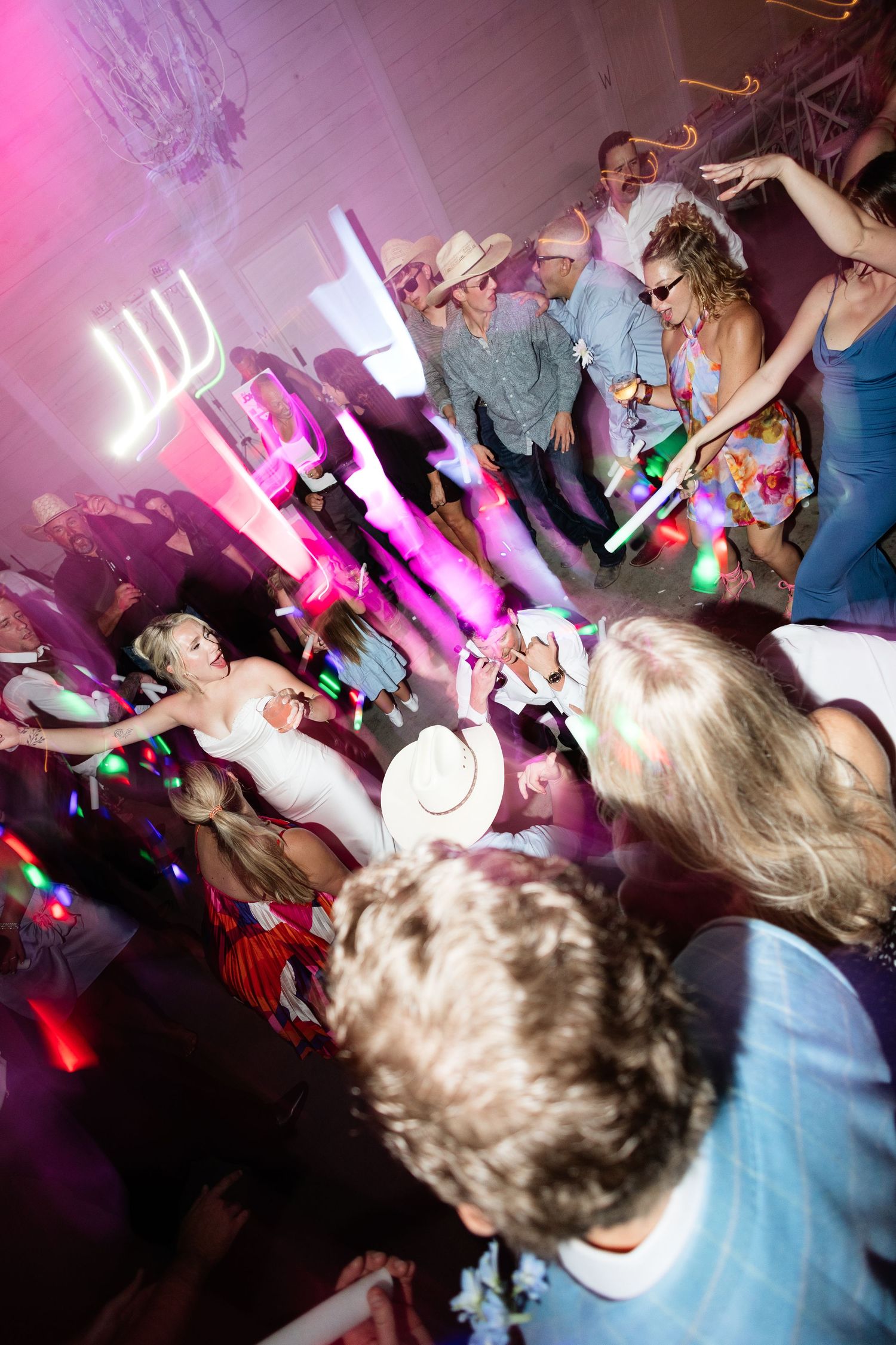 Lively wedding reception dance floor with guests in cowboy hats dancing under colorful pink and purple lights.