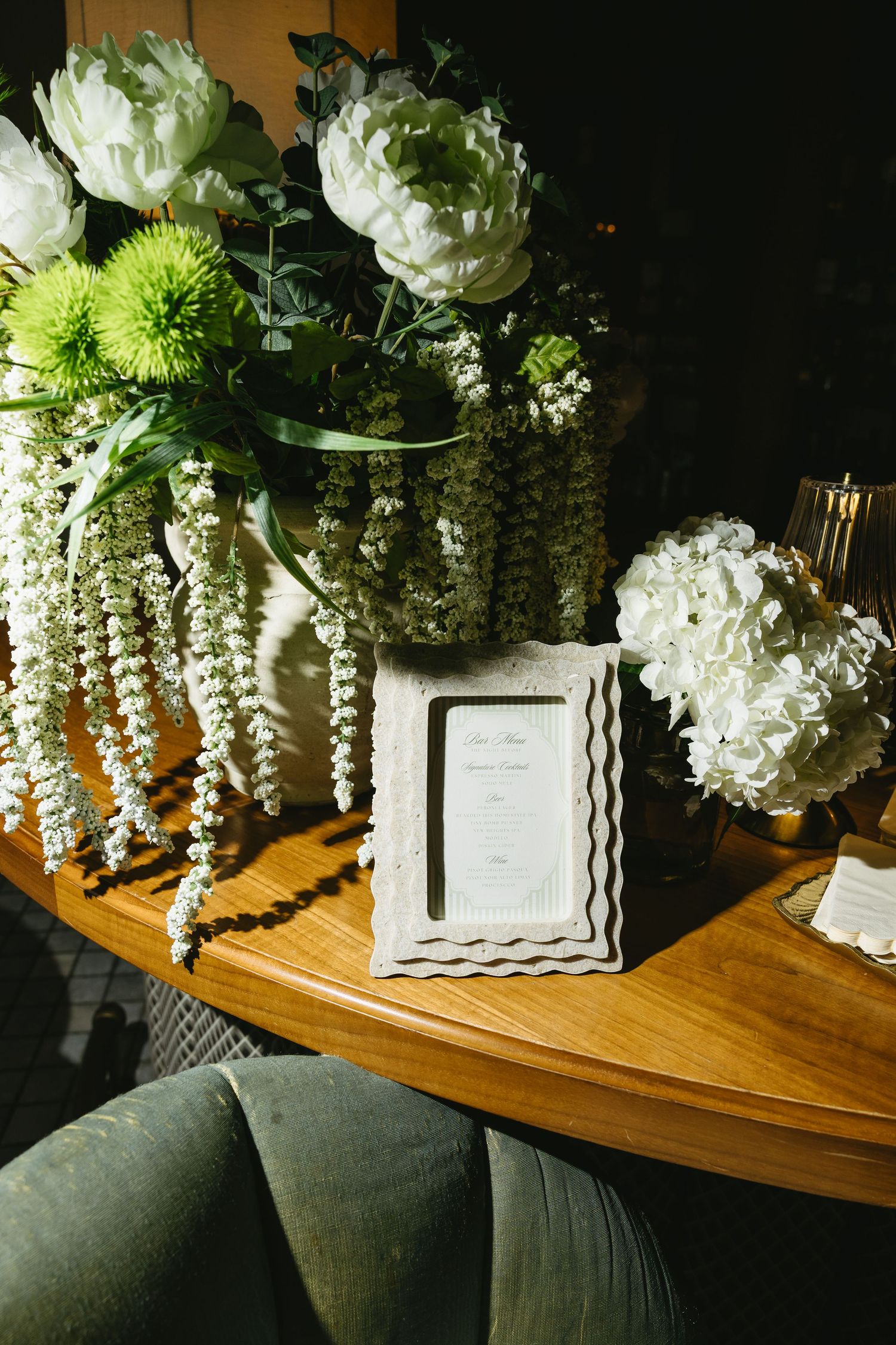 White and green floral arrangement with cascading elements sits on a polished wooden table near dark cushions.