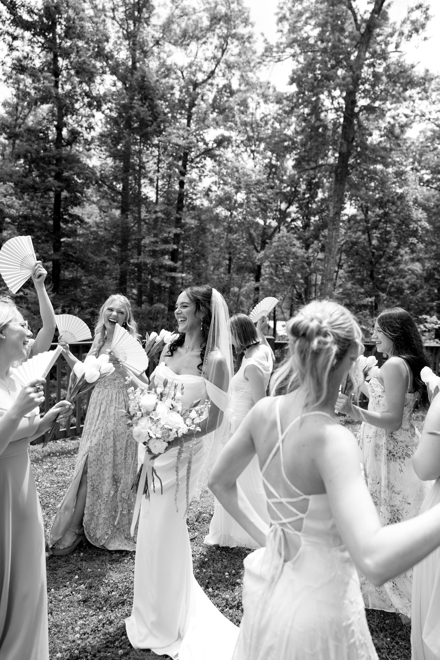Black and white photo of a bride holding a bouquet surrounded by bridesmaids with hand fans outdoors.