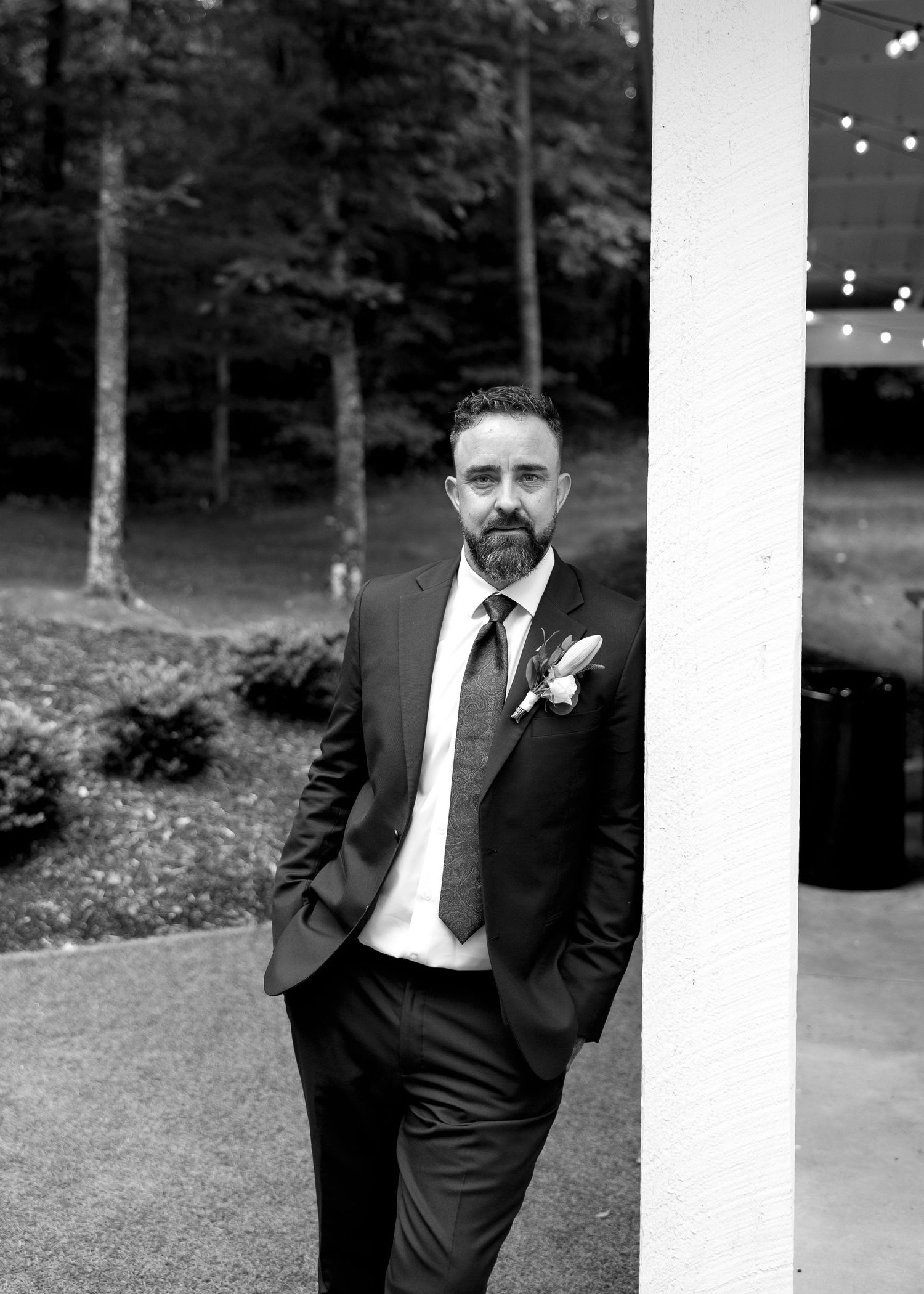 Groom in dark suit with boutonniere leans against white column at outdoor wedding venue surrounded by trees.