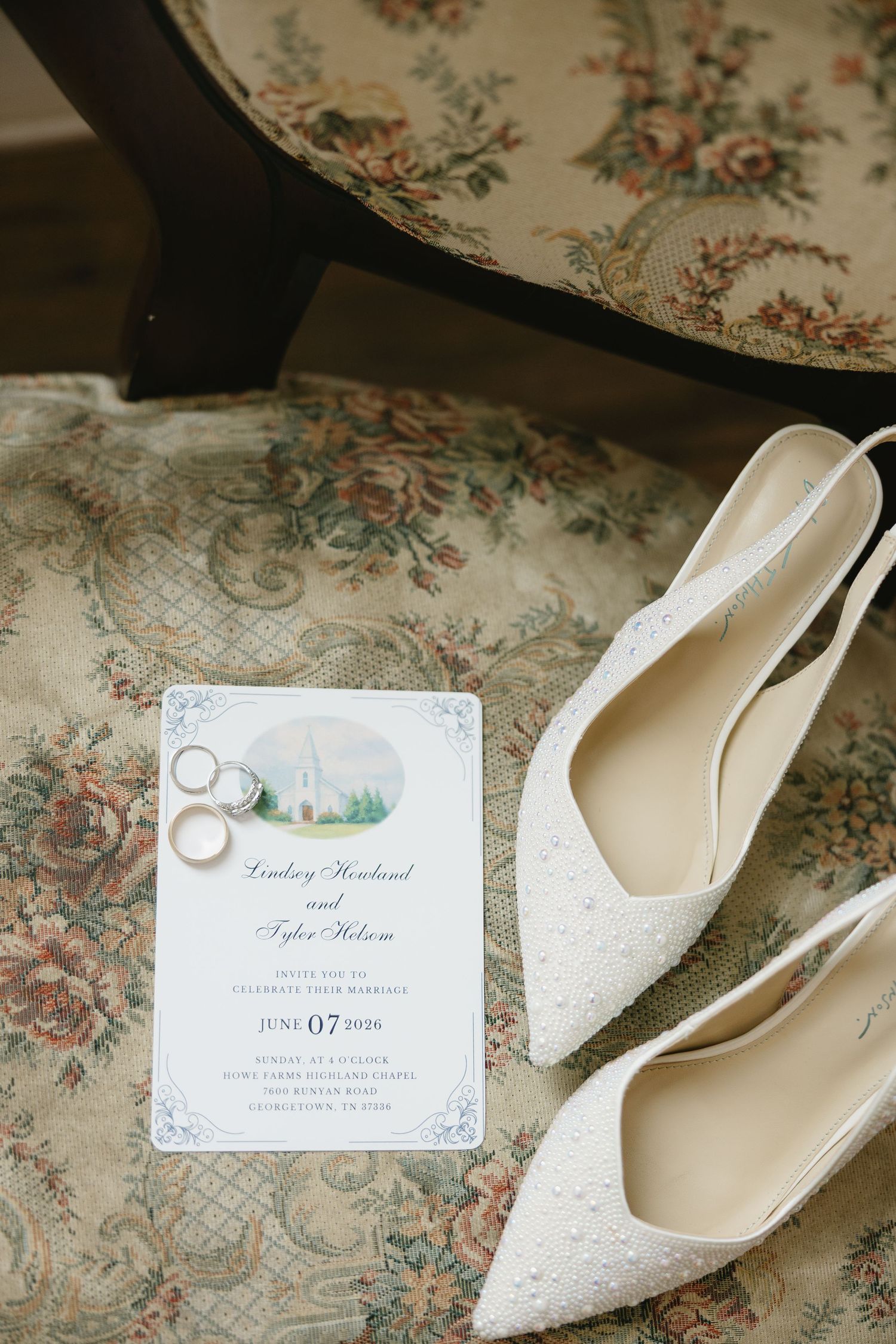 Wedding flat lay with lace heels, gold rings, and elegant invitation for Lindsey and Tyler on a floral chair.