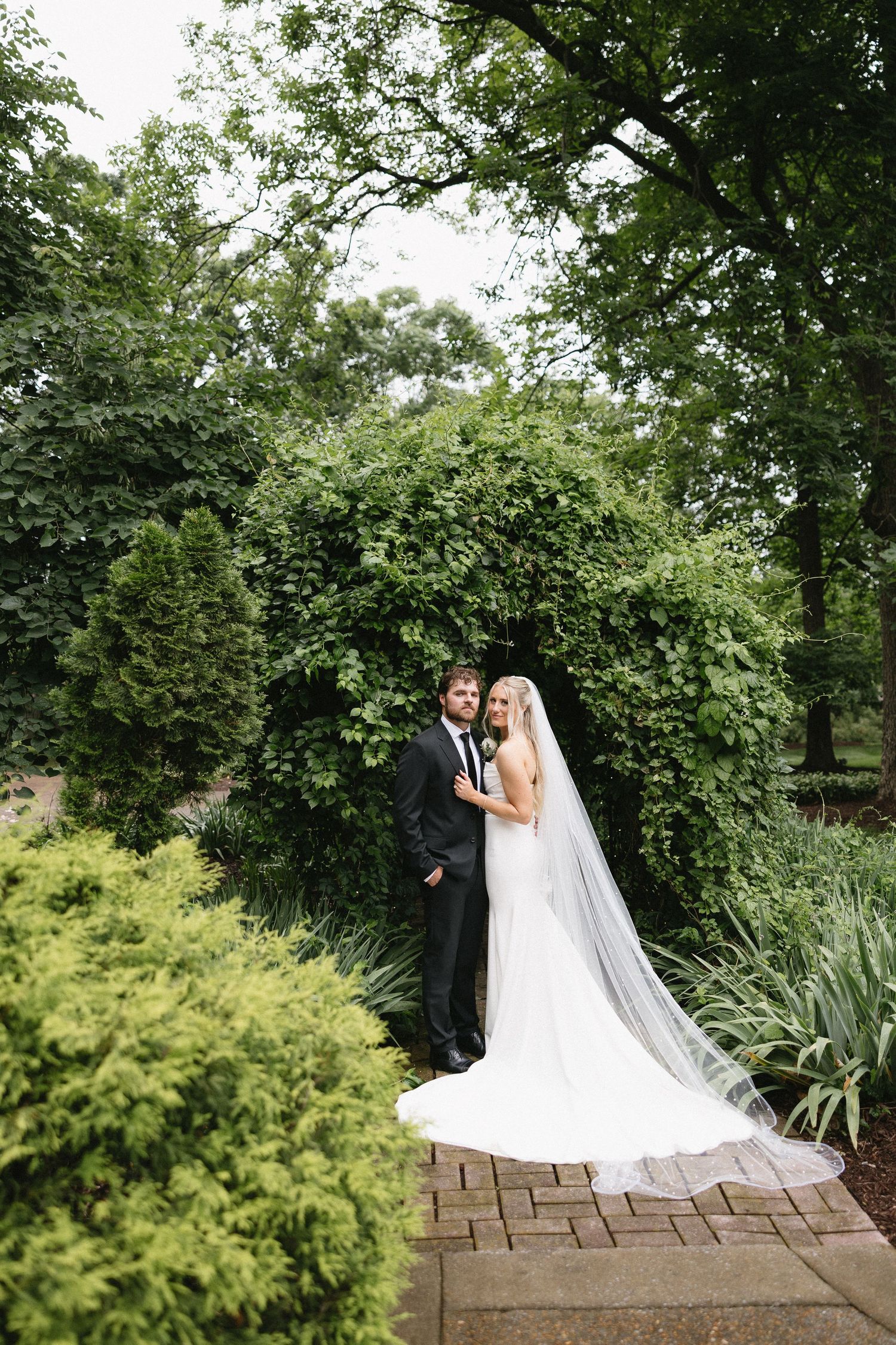 Bride and groom pose together in a lush garden setting, bride wearing a long veil and white gown.