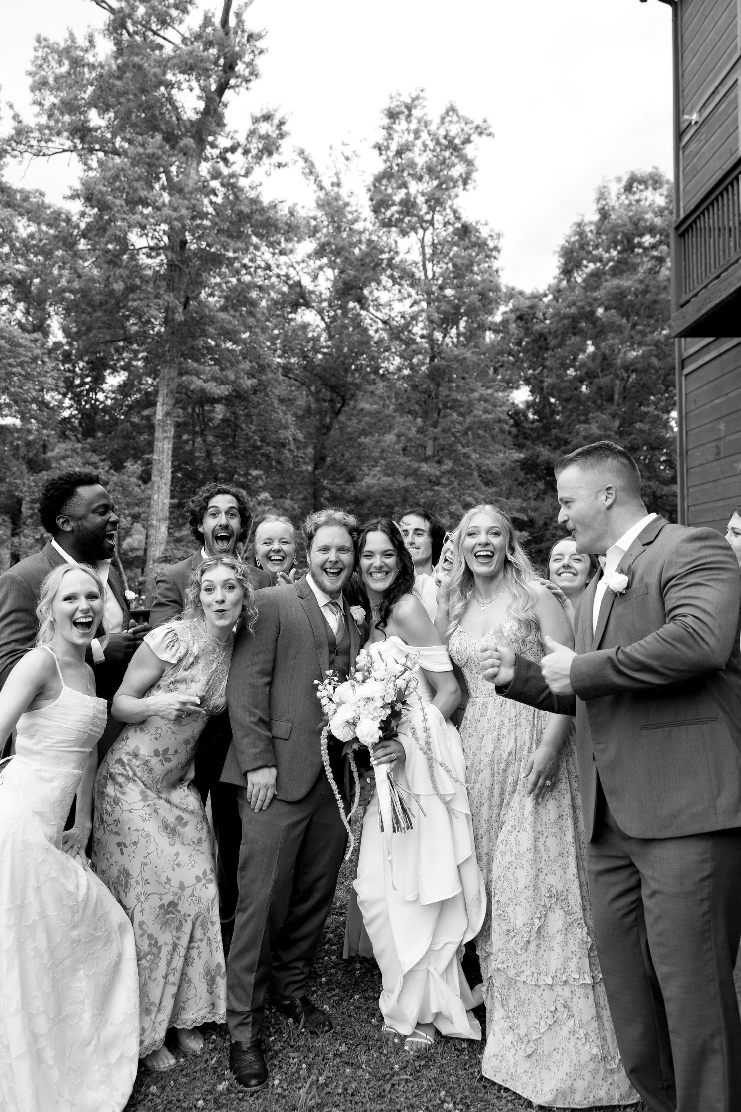 Black and white photo of a joyful wedding party laughing and celebrating outdoors with trees in the background.