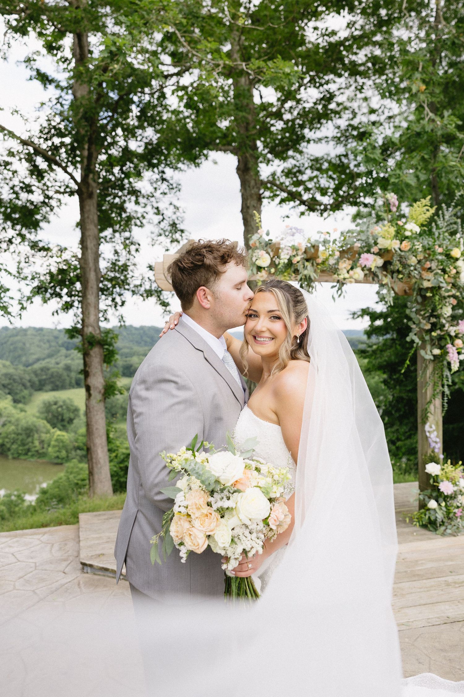 Groom in gray suit kisses bride wearing white gown and veil holding pastel bouquet at outdoor wedding ceremony.