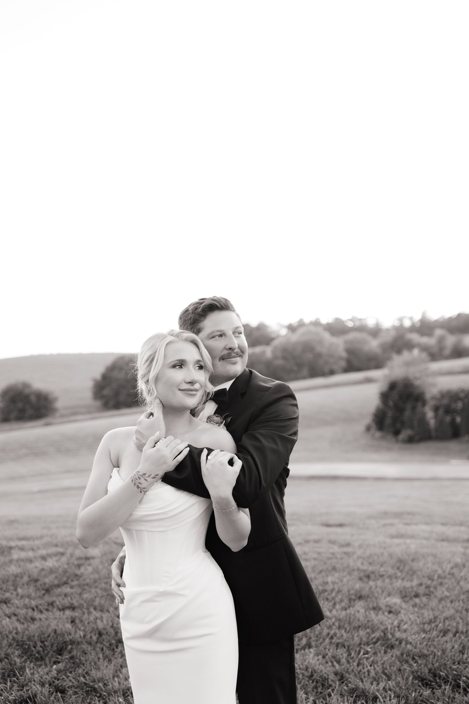 Black and white wedding photo of bride and groom embracing in a scenic field, groom holding bride from behind.