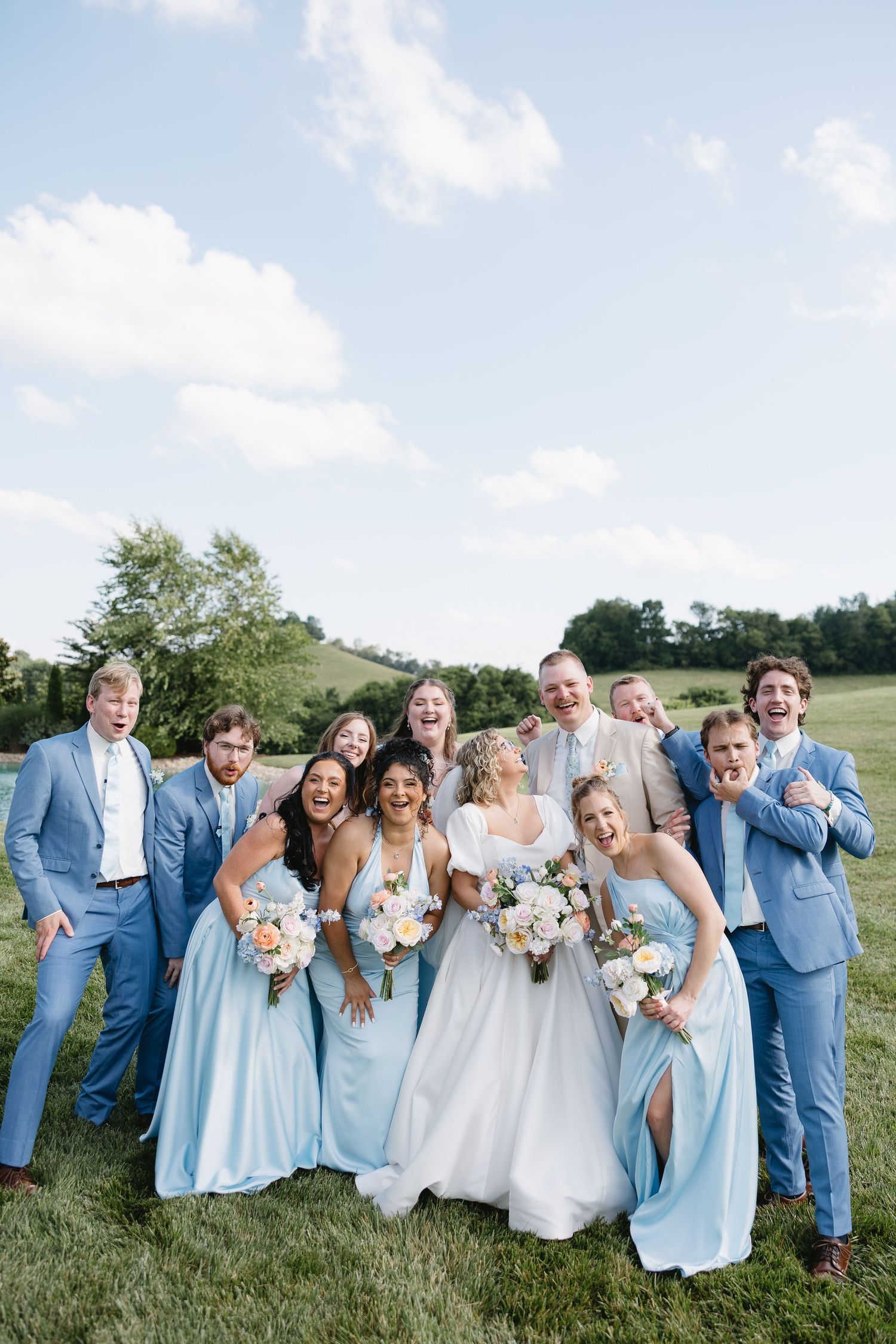 Bride and wedding party laughing outdoors in light blue dresses and suits on green lawn with scenic countryside background.