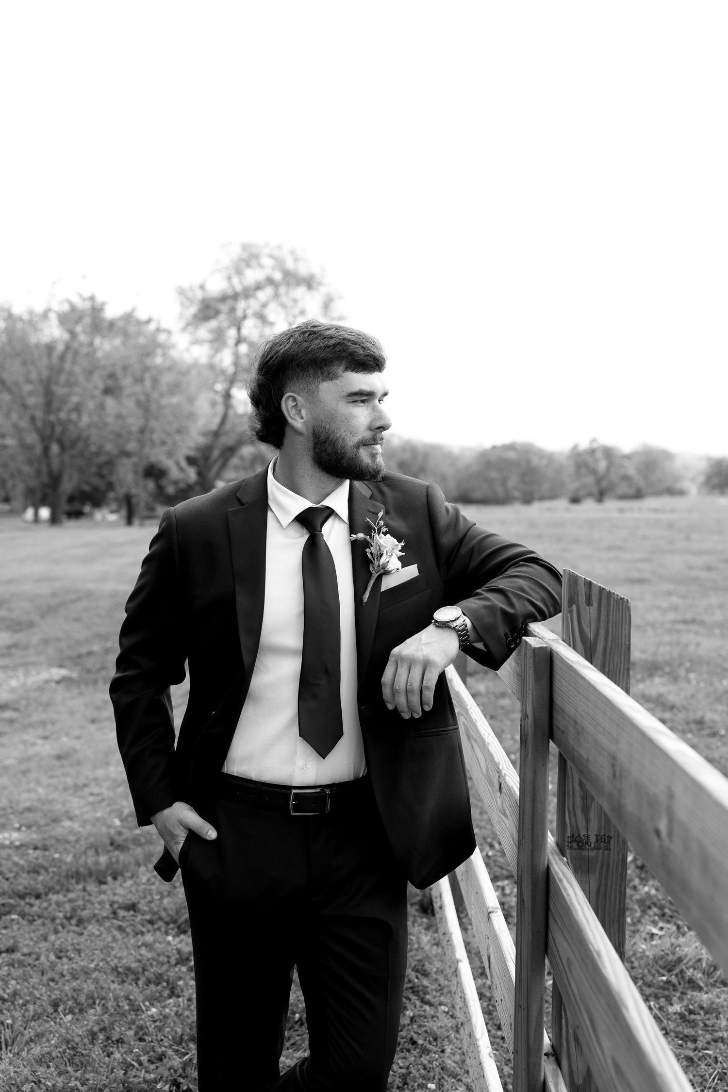 Black and white photo of a bearded groom in a suit with boutonniere leaning on a wooden fence in a rural field.