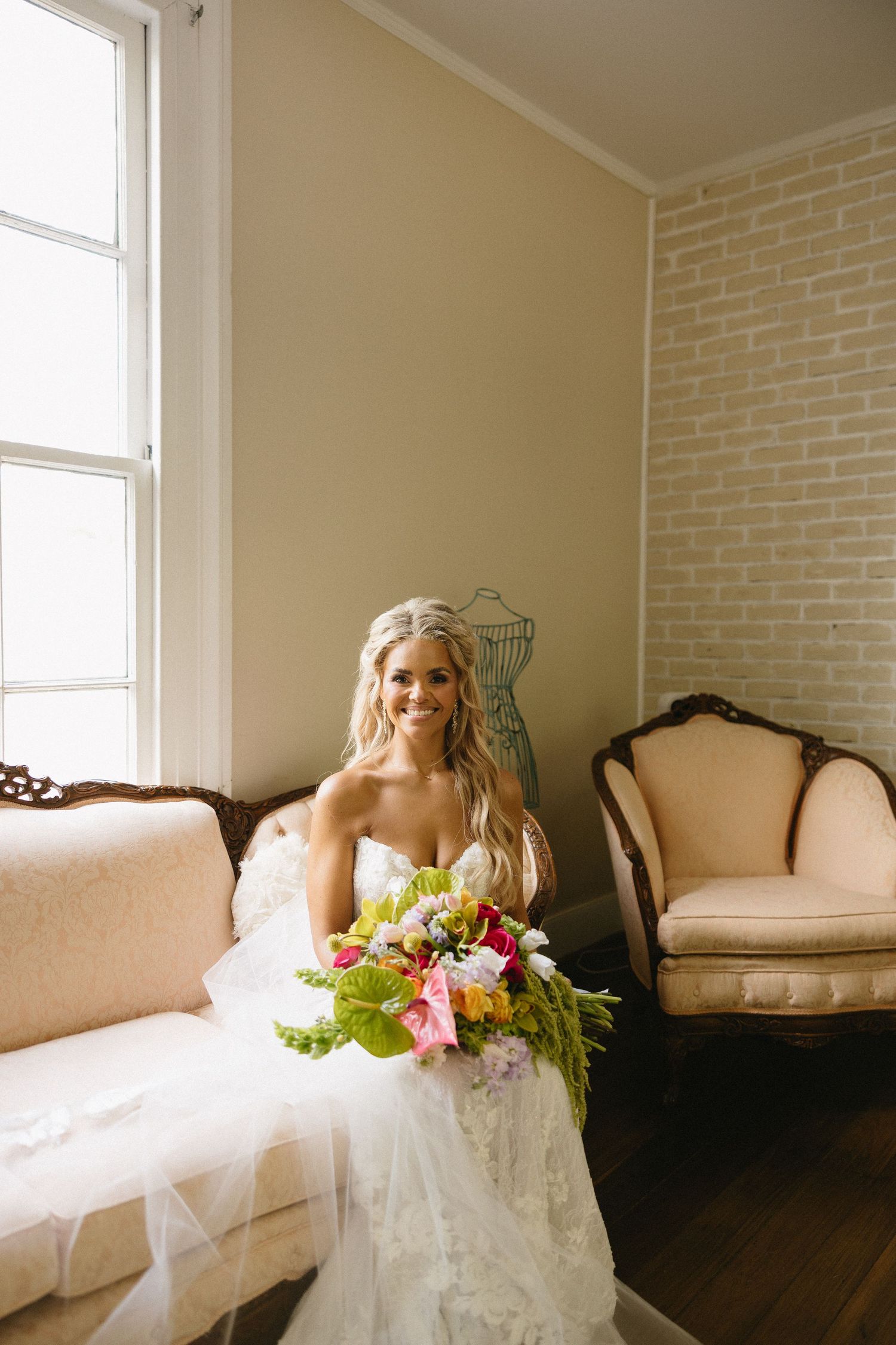 Smiling bride in white lace gown holding colorful bouquet seated on vintage sofa near window in elegant room.