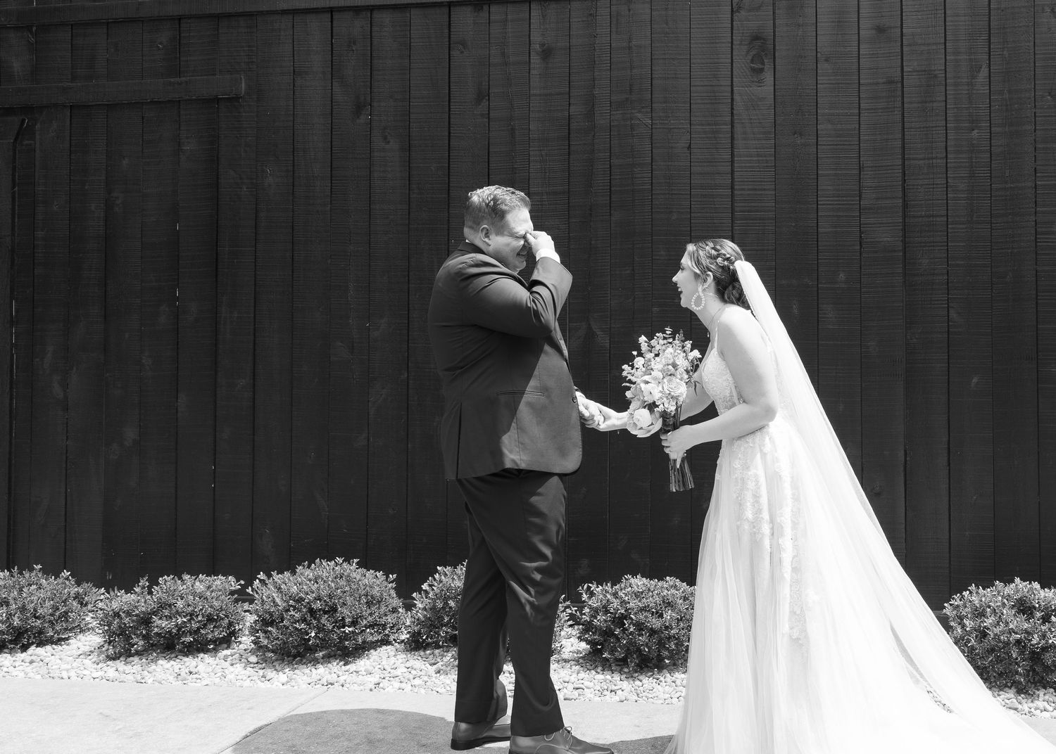 Black and white photo of a groom wiping tears during first look with bride holding bouquet at barn wedding.
