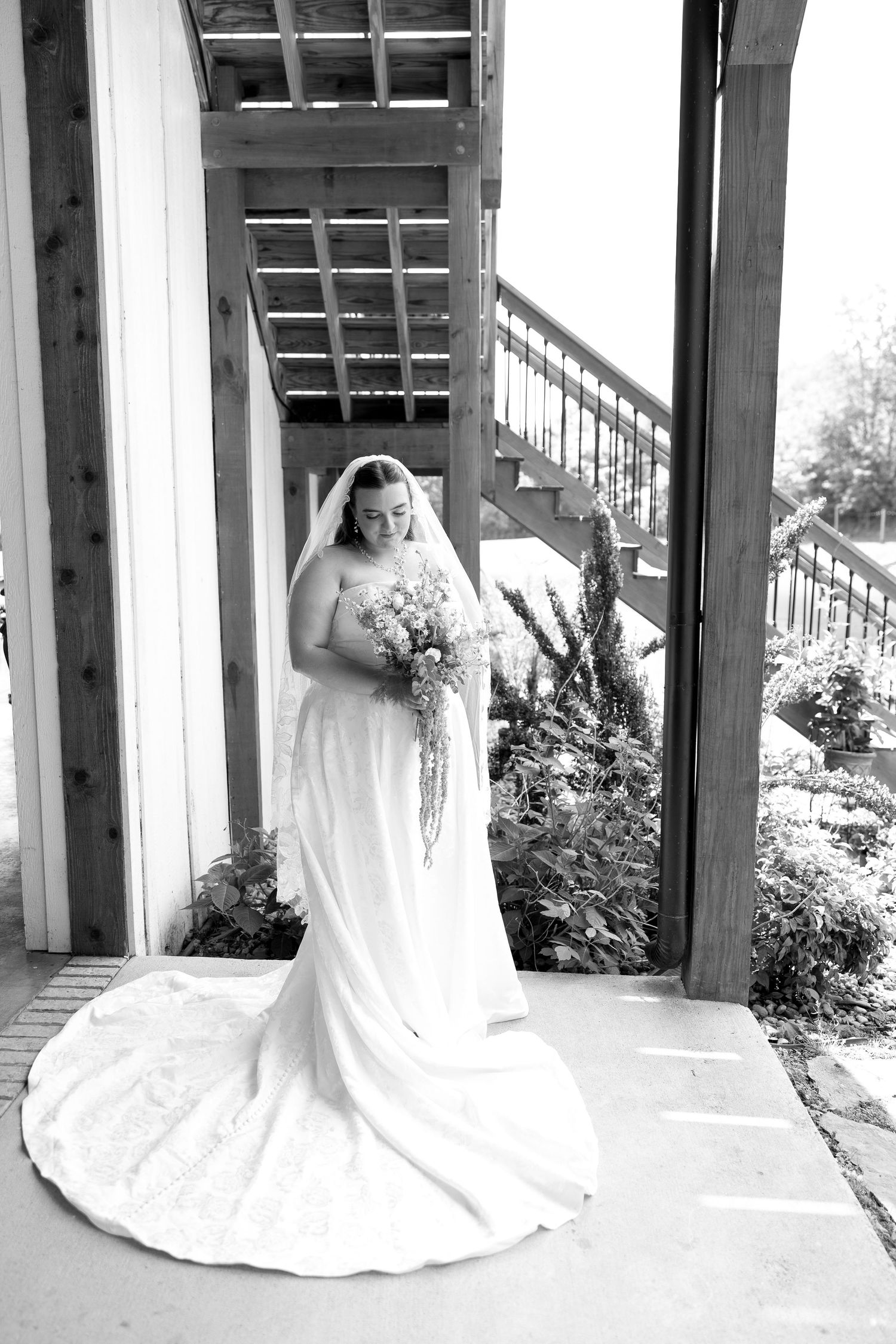 Black and white bridal portrait of a bride in a white gown holding a bouquet, standing under a wooden pergola.
