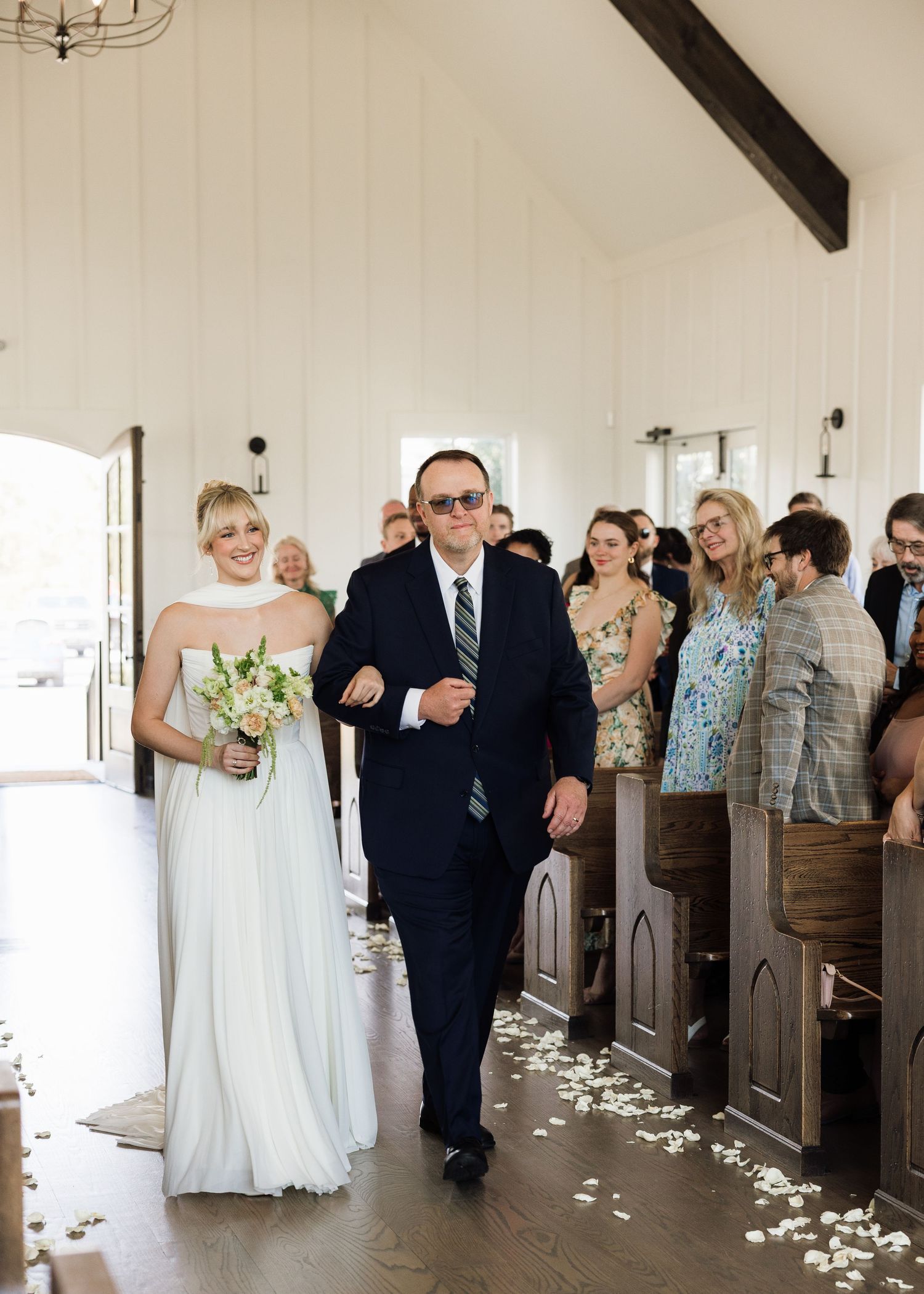 Bride in white gown walks down the aisle with escort in navy suit at elegant chapel wedding ceremony.