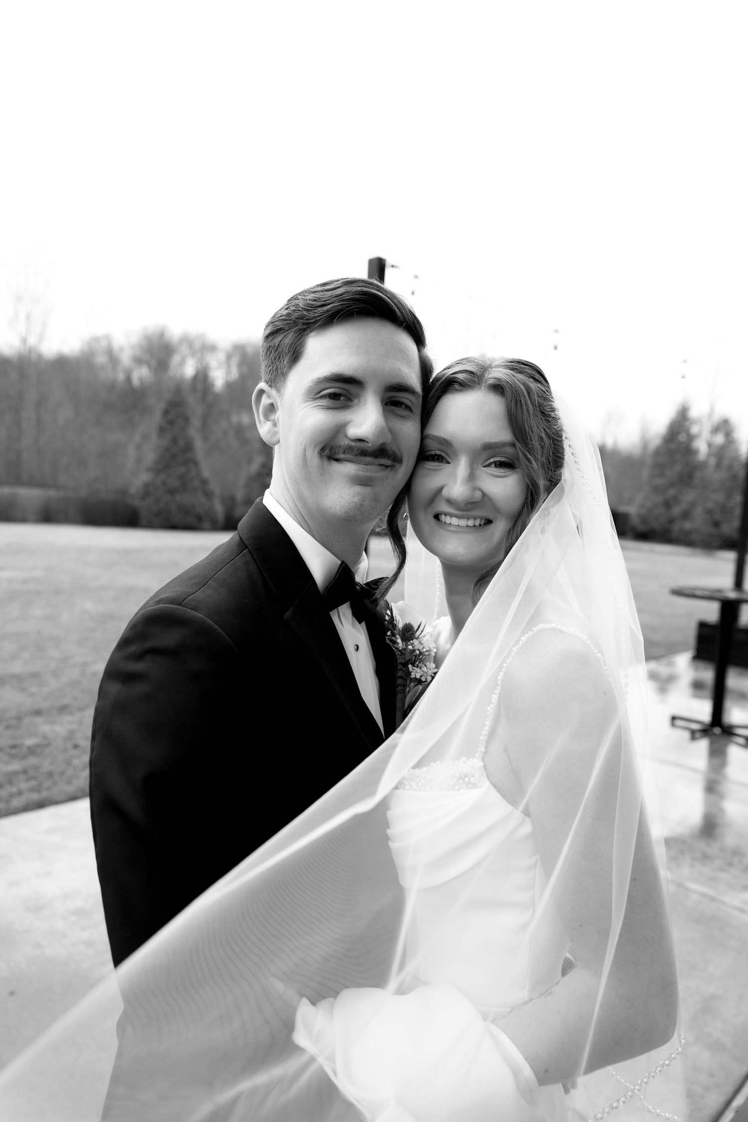 Black and white wedding portrait of smiling bride in veil and groom in tuxedo embracing outdoors.