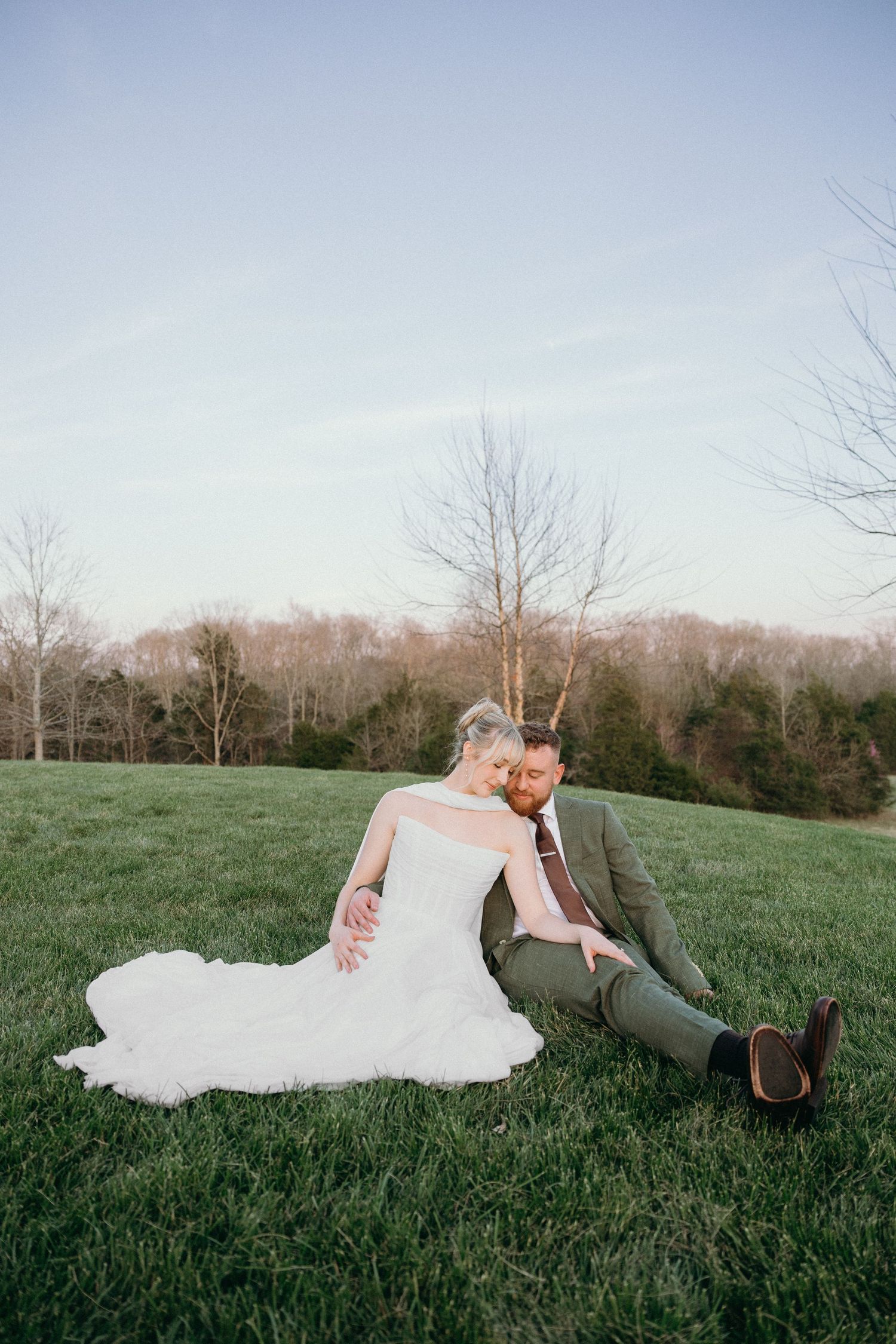 Bride and groom sitting together on lush green grass at sunset, surrounded by bare winter trees.