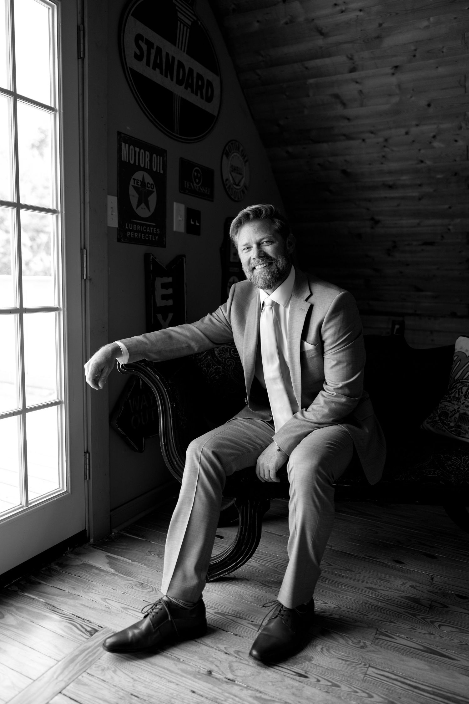 Black and white portrait of a bearded man in a suit sitting confidently in a chair beside a window in a rustic room.