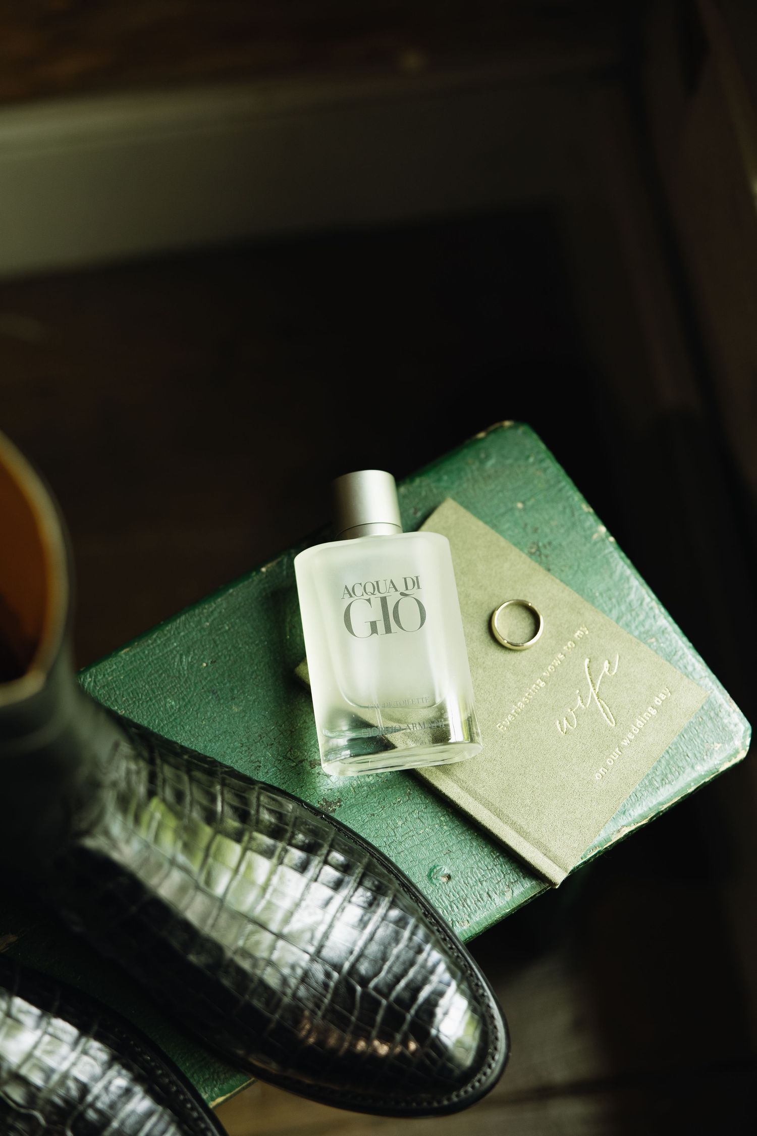 Acqua Di Gio cologne bottle resting on green book with gold wedding ring and dress shoes nearby.