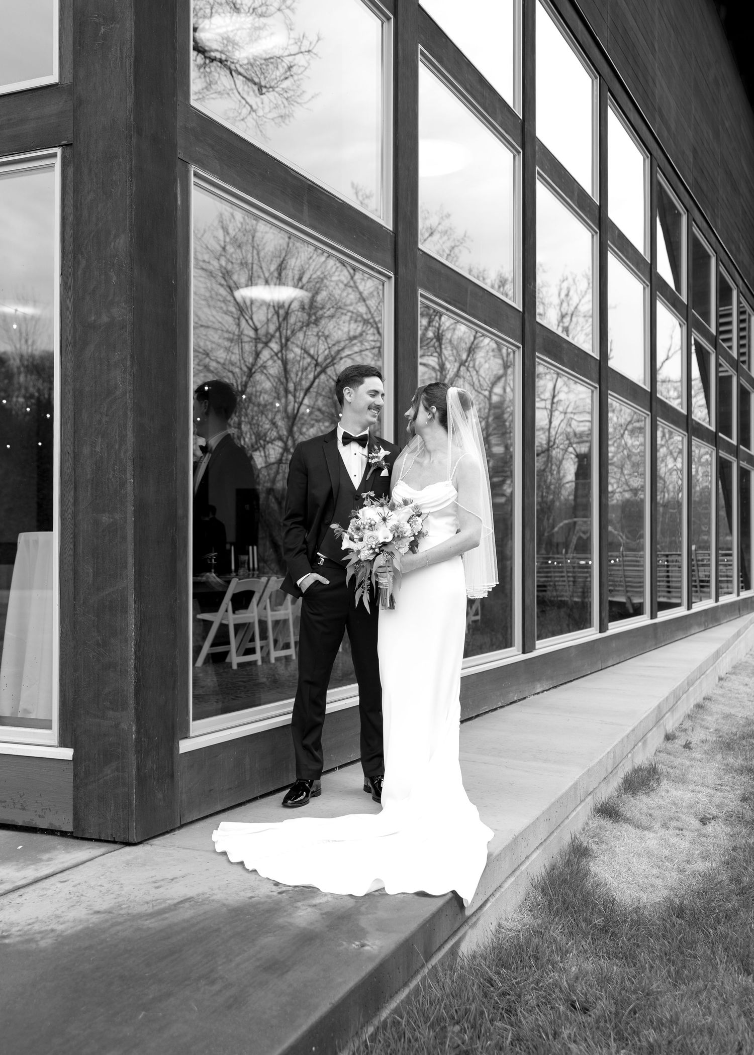 Black and white photo of bride and groom standing outside modern building with large windows.