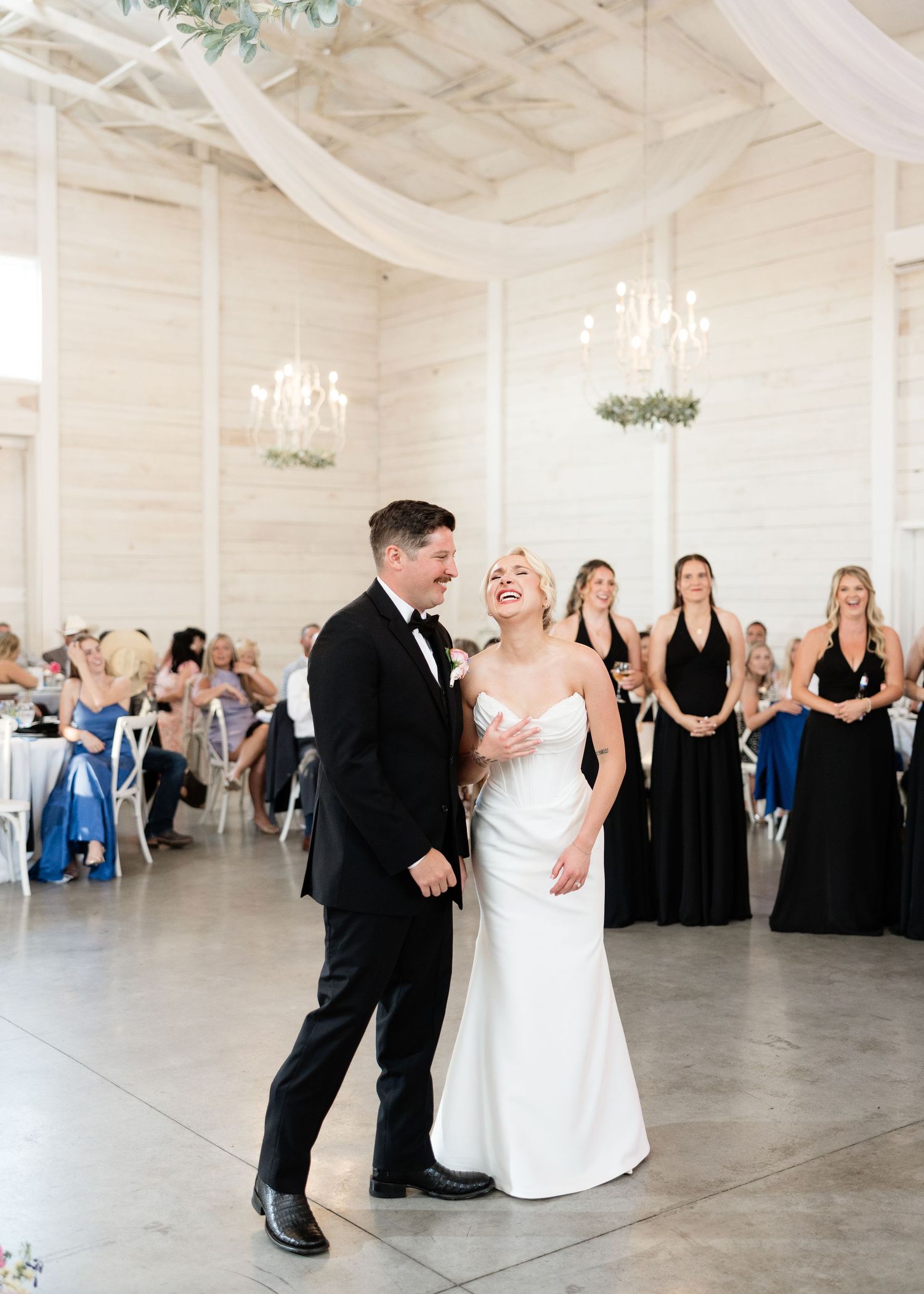 Bride and groom share first dance in elegant white reception hall, surrounded by bridesmaids in black gowns.