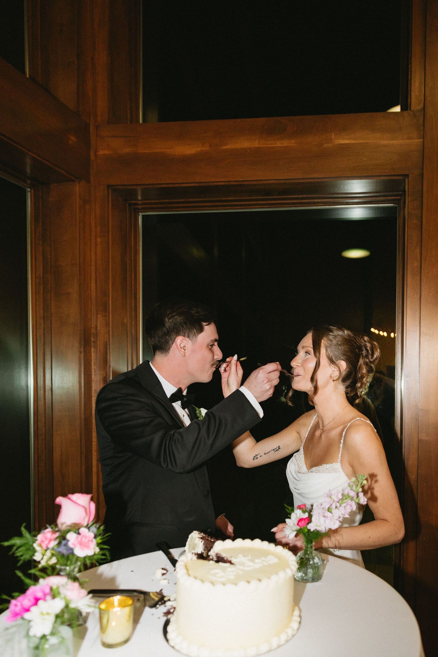 Bride and groom sharing wedding cake at reception table with pink flowers and candlelight in elegant wood-paneled venue.