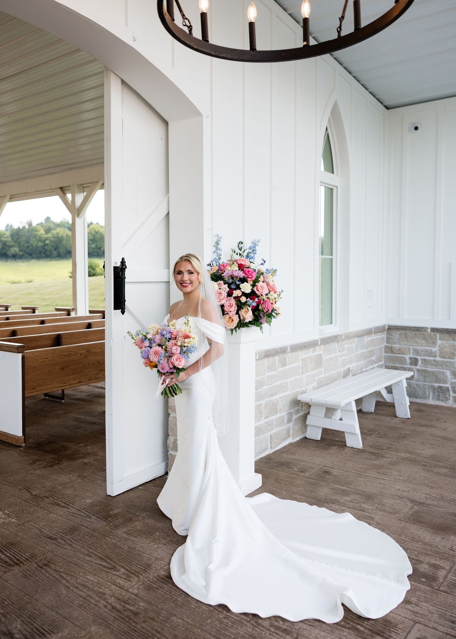 Bride in white mermaid gown holds colorful bouquet inside elegant white chapel with wooden pews and chandelier.