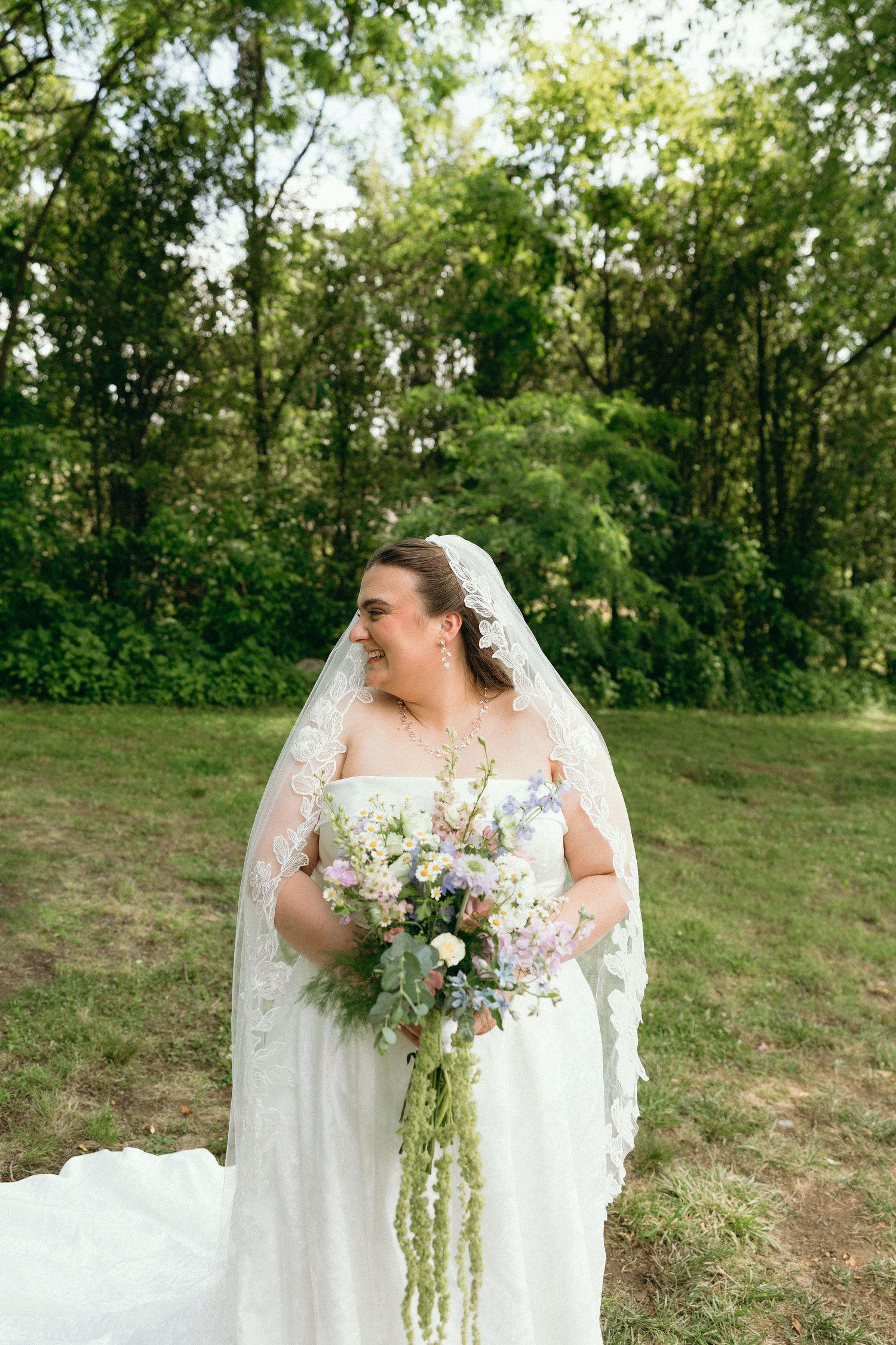 Smiling bride in lace veil holding cascading wildflower bouquet, standing outdoors in green garden setting.