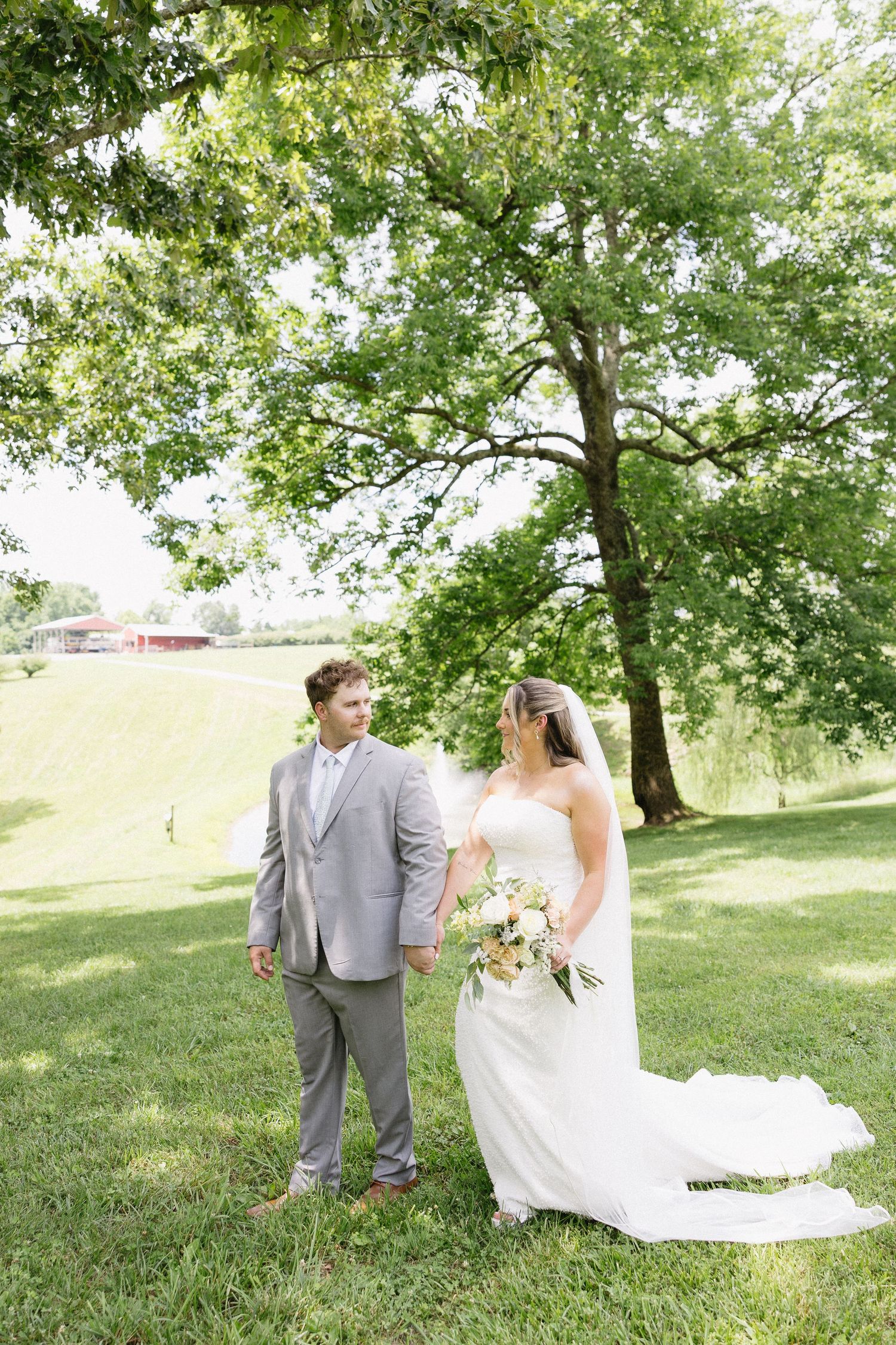 Bride and groom stand under a large oak tree on lush green grass, bride holding a bouquet in a white gown.