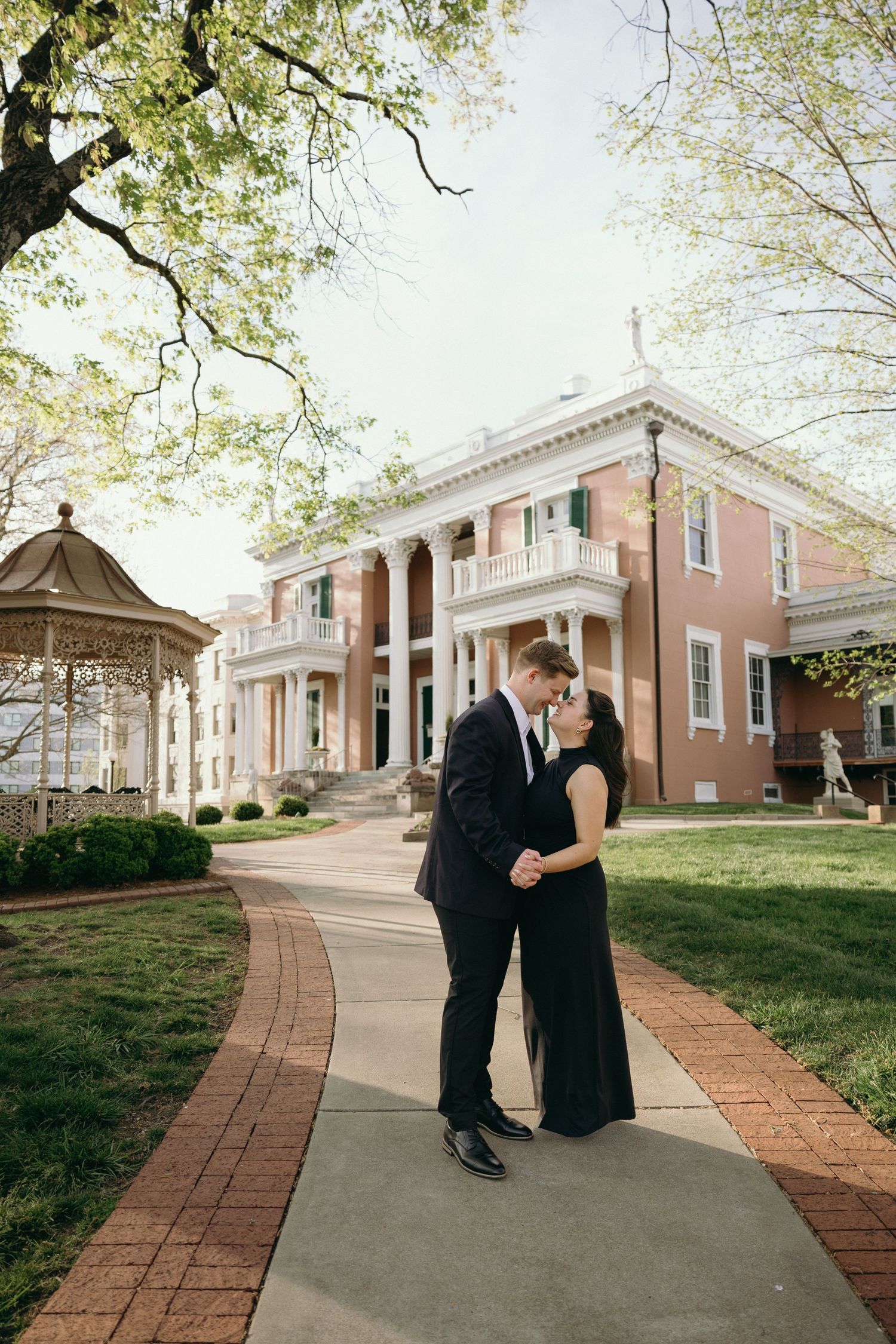 Couple sharing a romantic kiss in formal attire on a brick pathway outside an elegant Southern mansion in spring.