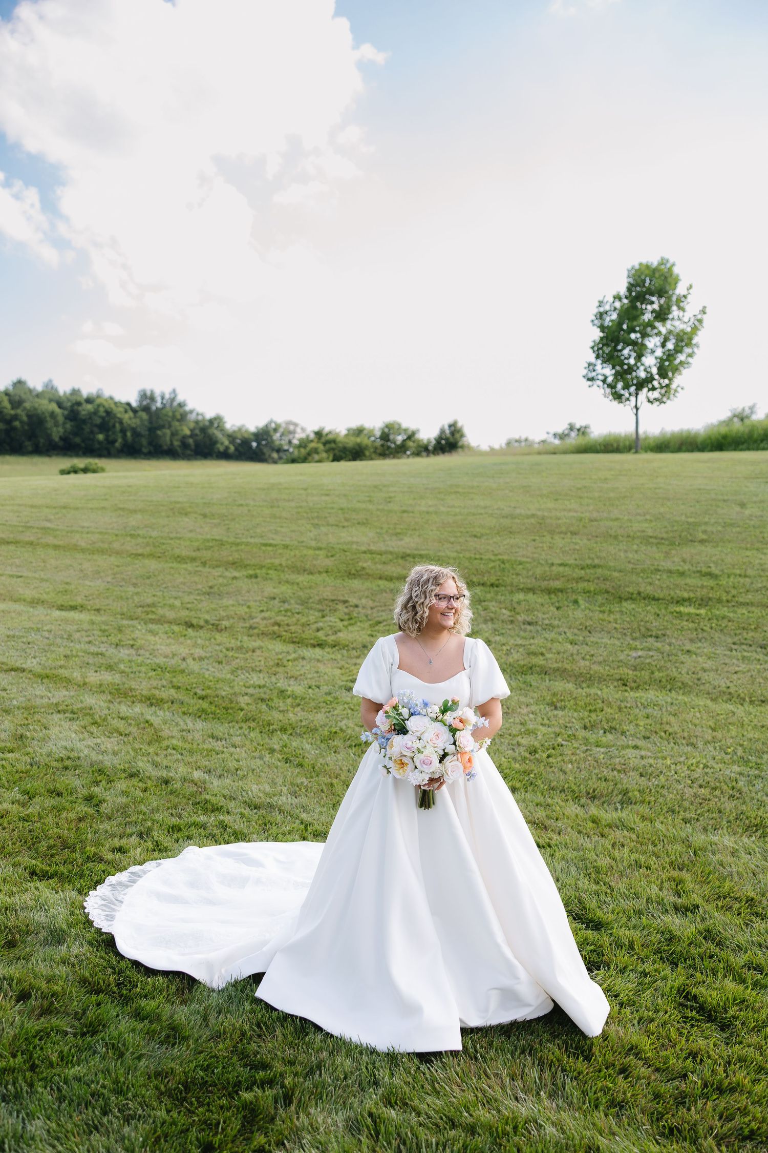 Bride in white ball gown with train holds floral bouquet on lush green lawn under blue sky.