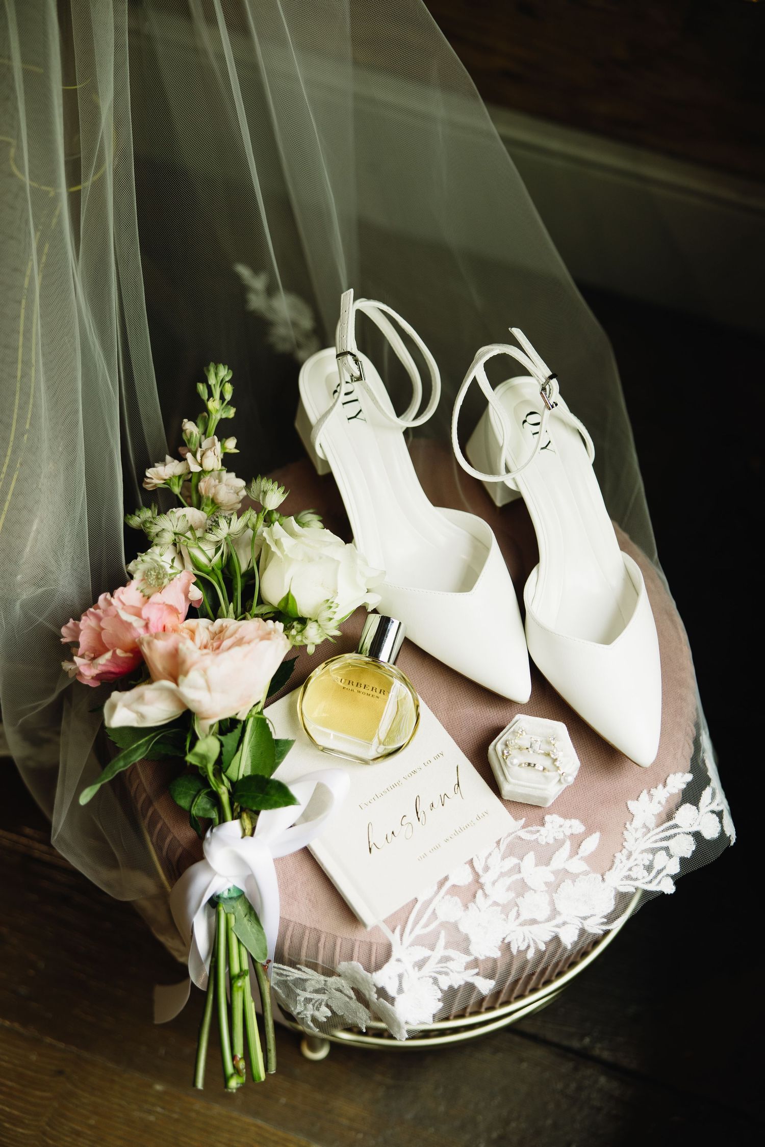 Elegant bridal flat lay with white slingback heels, pink bouquet, perfume bottle, ring box, and lace veil on a tray.