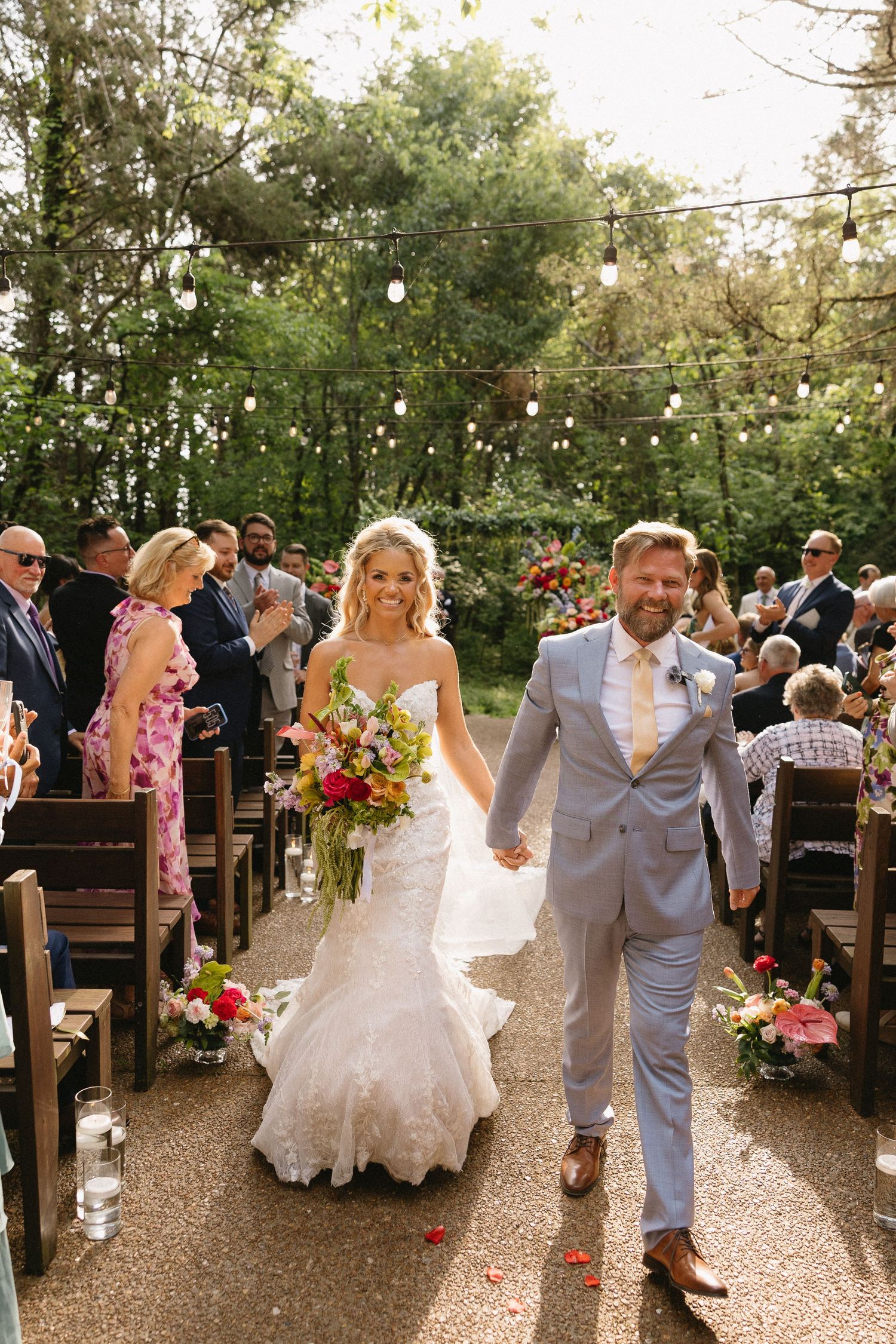 Bride and groom walk down outdoor aisle hand-in-hand, she holds colorful bouquet, guests applaud in sunlit garden ceremony.