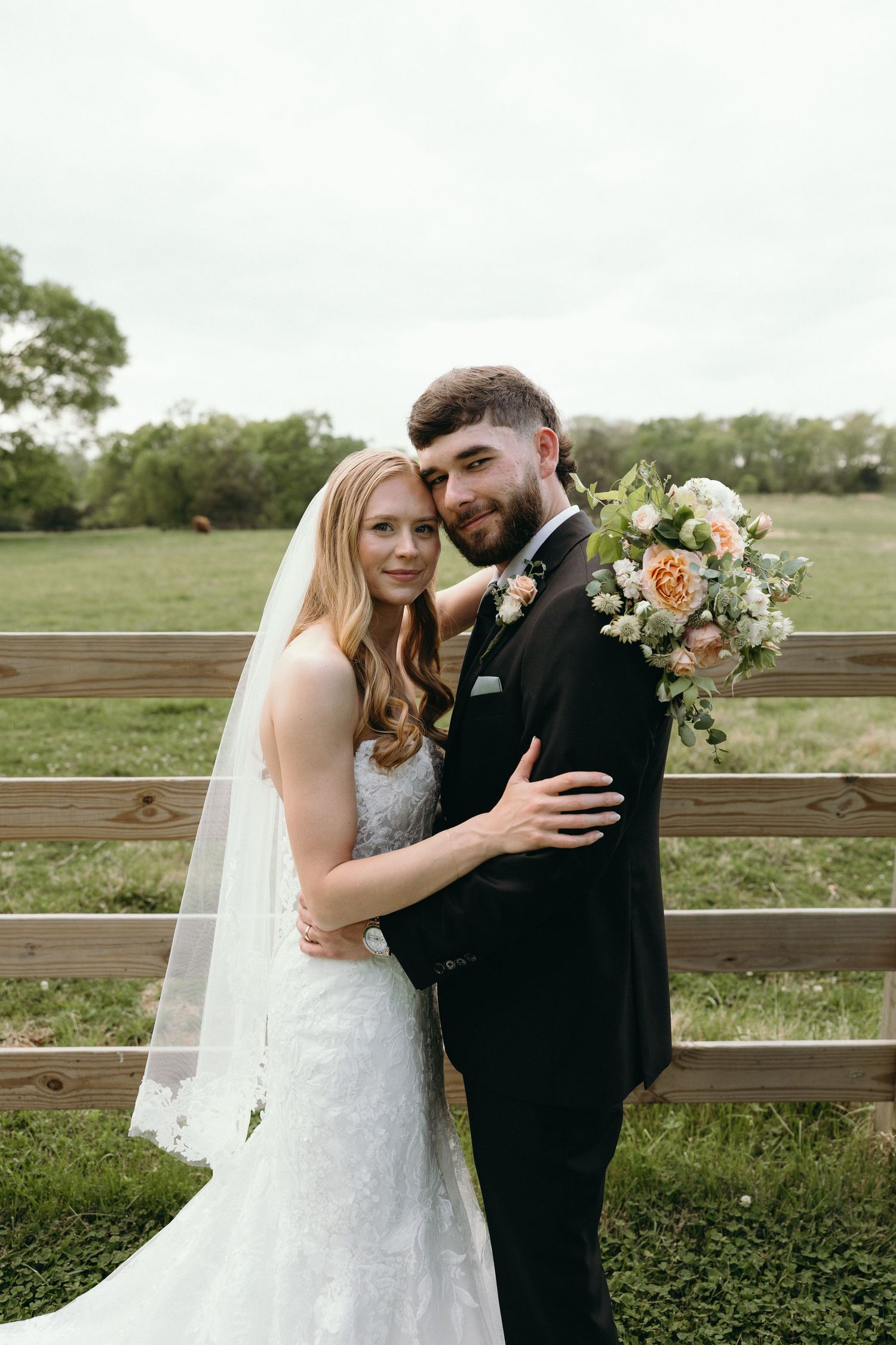Bride and groom embrace in front of a rustic wooden fence at an outdoor farm wedding, bride holding a peach bouquet.