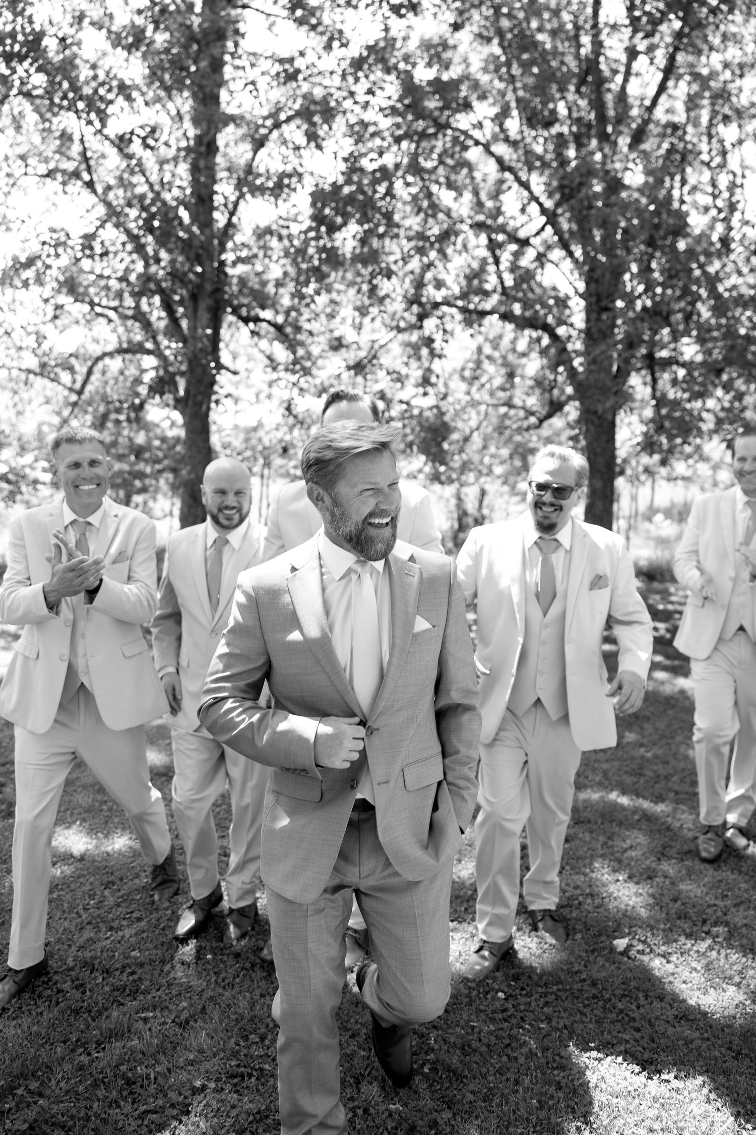 Black and white photo of groom laughing with groomsmen in light suits outdoors among trees at a wedding.