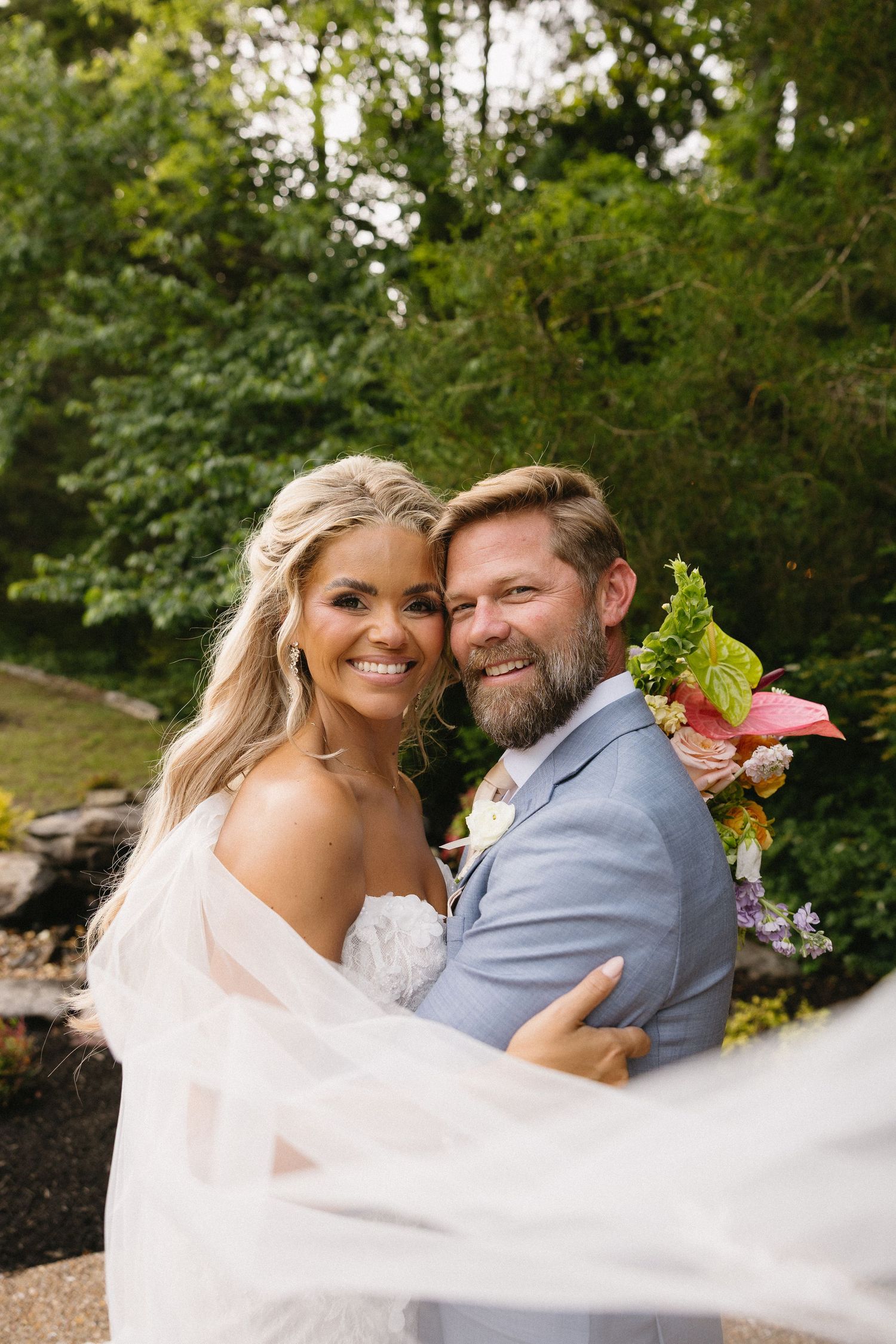 Smiling bride and groom embrace outdoors, bride in strapless gown with veil, groom in blue suit with floral boutonniere.