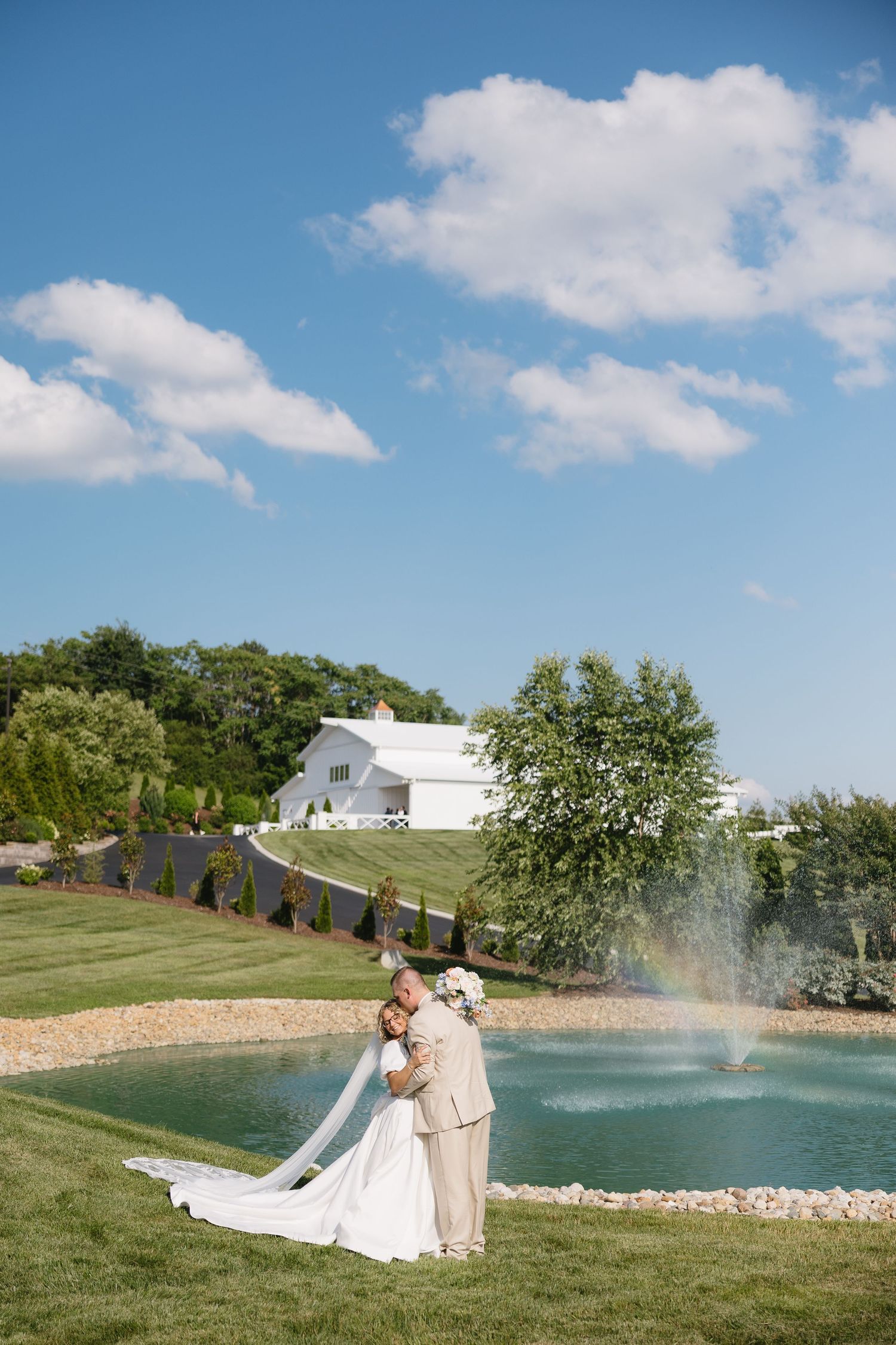 Bride and groom embrace by a pond with fountain, white barn venue and blue sky in background.