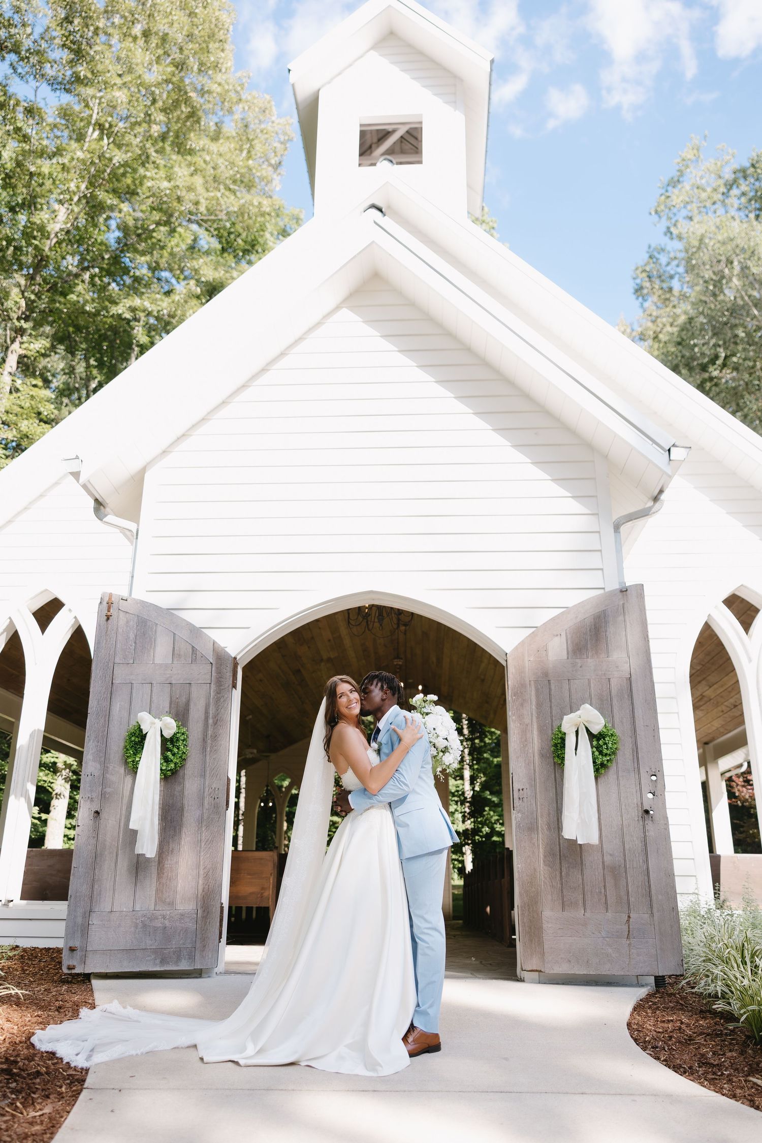 Bride and groom embrace at white chapel doors adorned with greenery wreaths on sunny wedding day.
