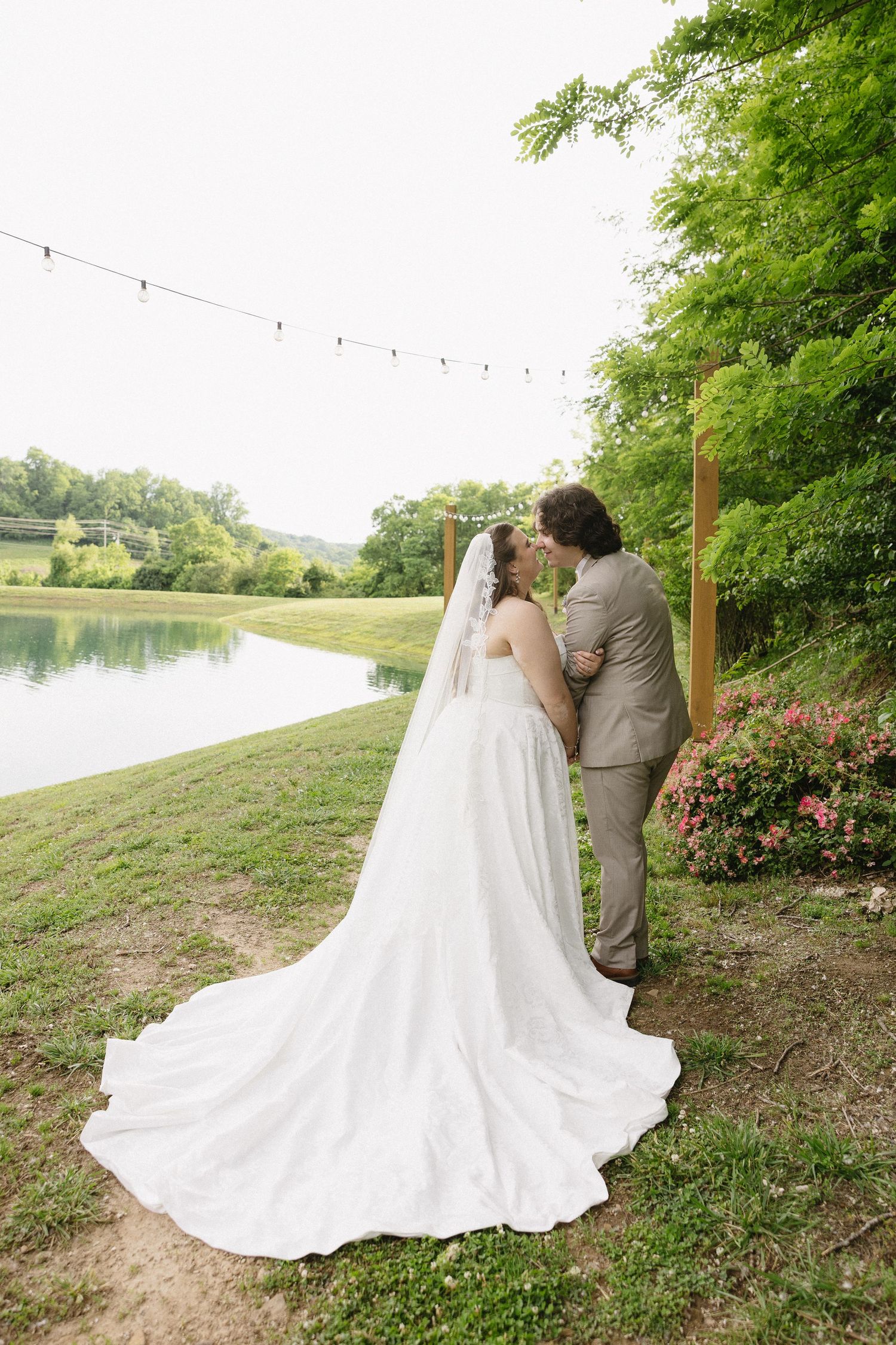 Bride and groom share a romantic moment by a serene pond, surrounded by lush greenery and pink flowers at an outdoor wedding.