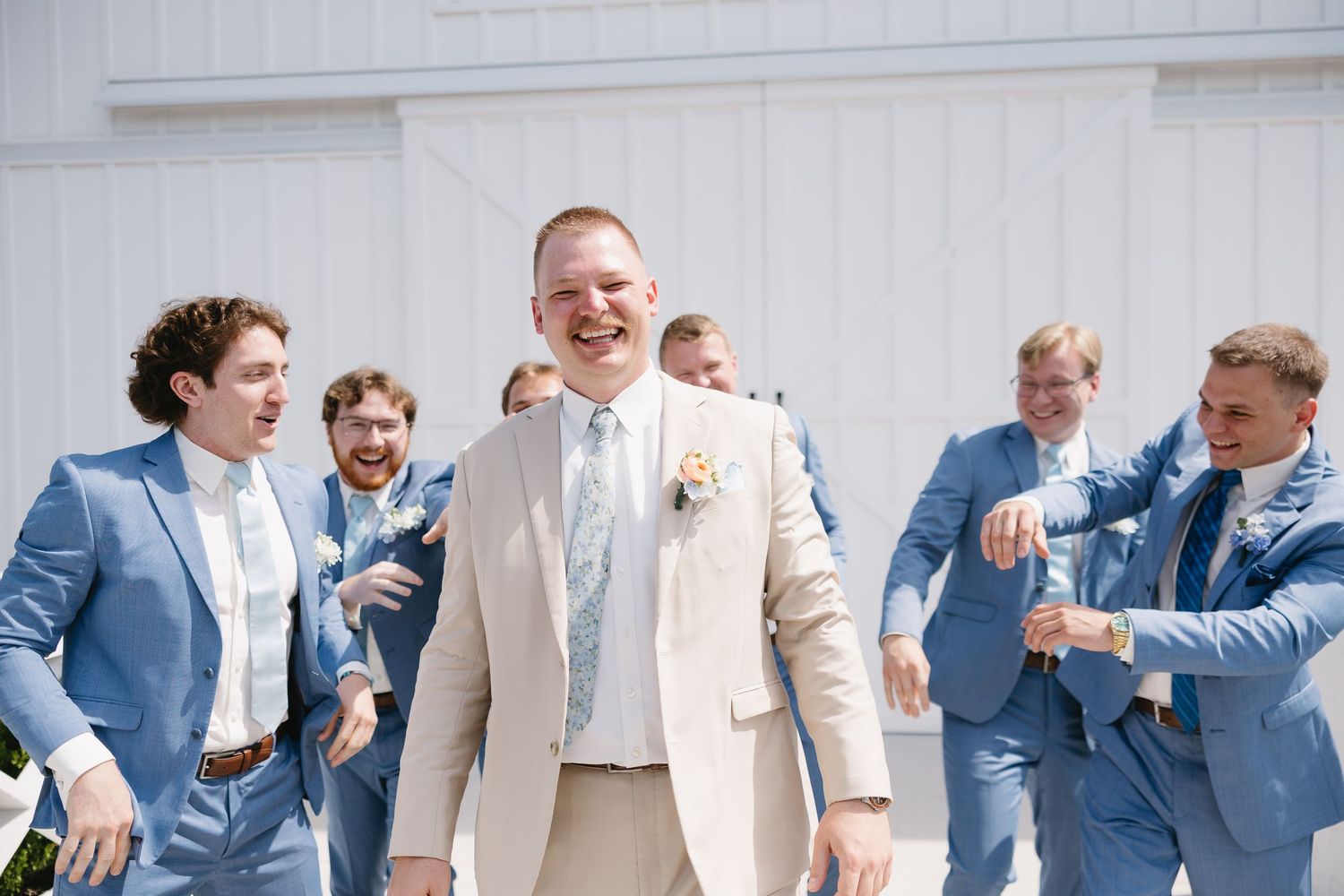 Groom in beige suit laughing with groomsmen in blue suits outside a white barn at a wedding.
